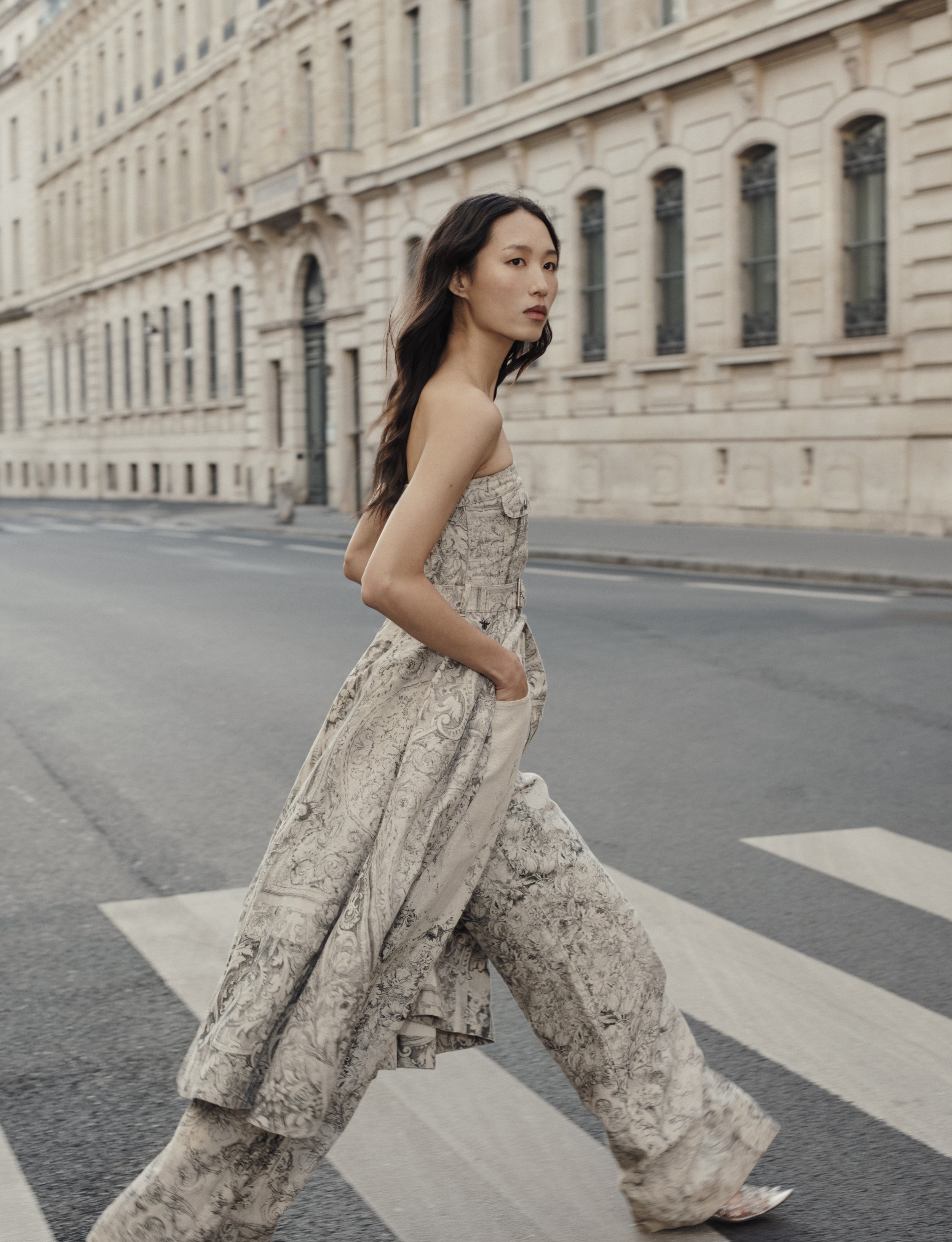 Une femme marche sur un passage piéton dans une rue urbaine, portant une tenue élégante beige avec des motifs.