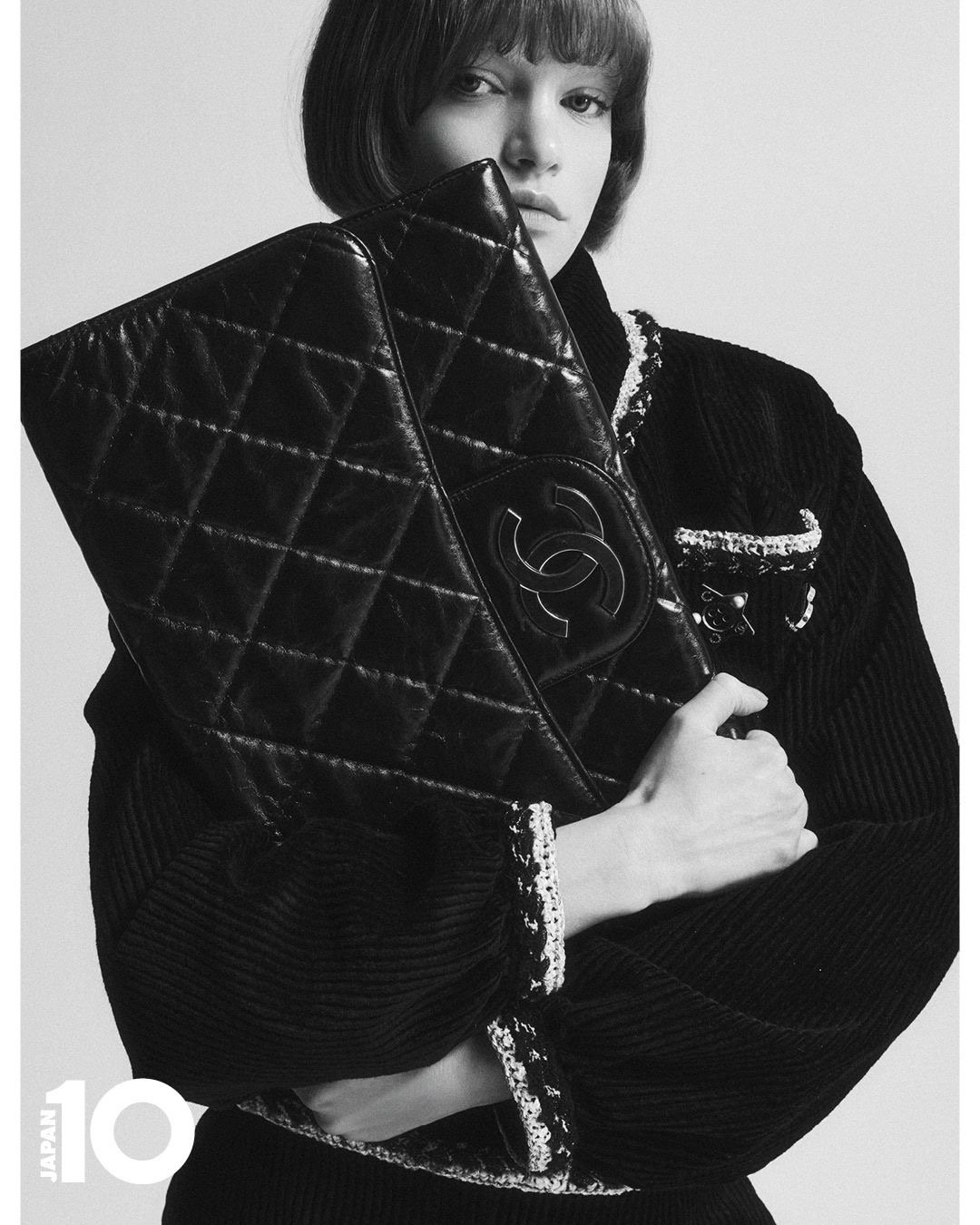 Une femme avec une coiffure courte, portant un vêtement en tissu noir, tient un sac à main en cuir noir à motif matelassé avec le logo de Chanel, dans une pose élégante. Image en noir et blanc avec texte 'JAPAN 10' en bas à gauche.