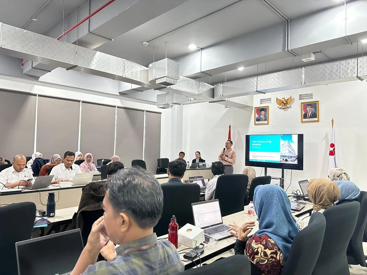 A conference room with people attending a presentation. The presenter is standing next to a screen displaying a slide. Attendees are seated at tables with laptops and notebooks, some of them wearing headscarves. The room has framed portraits on the wall, along with the national emblem and flags.