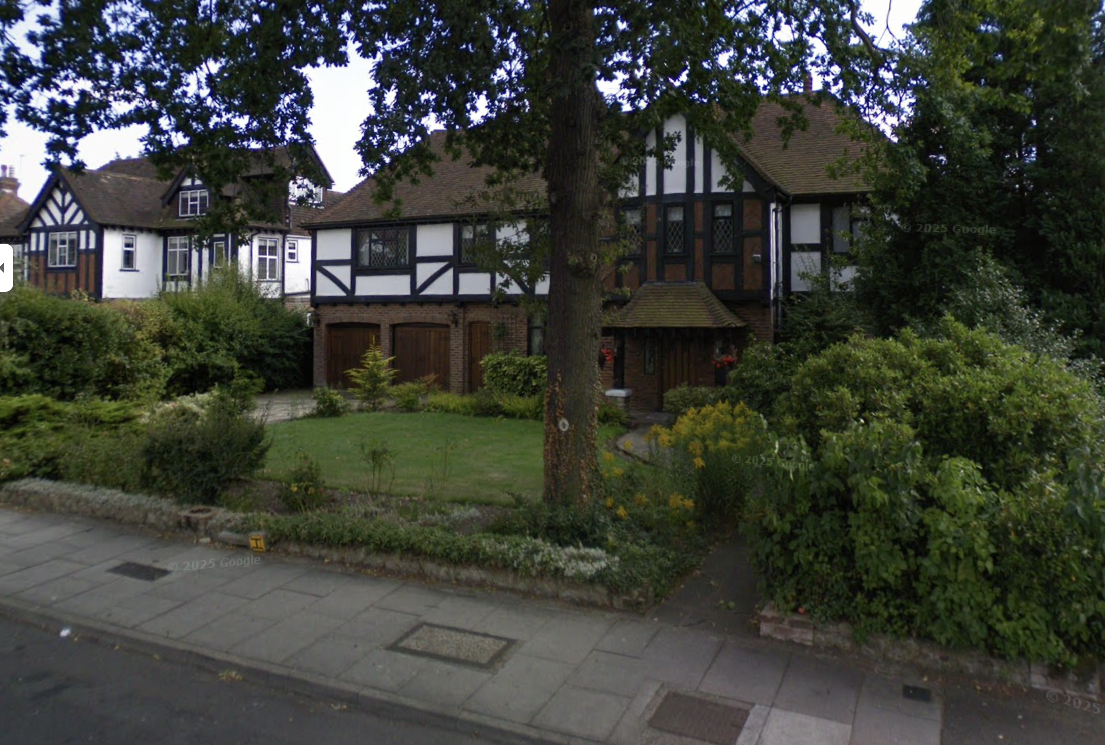 Large Tudor-style house with white and black timber framing, a brick foundation, and a sloped roof, surrounded by green bushes, trees, and a manicured lawn in a residential neighborhood.