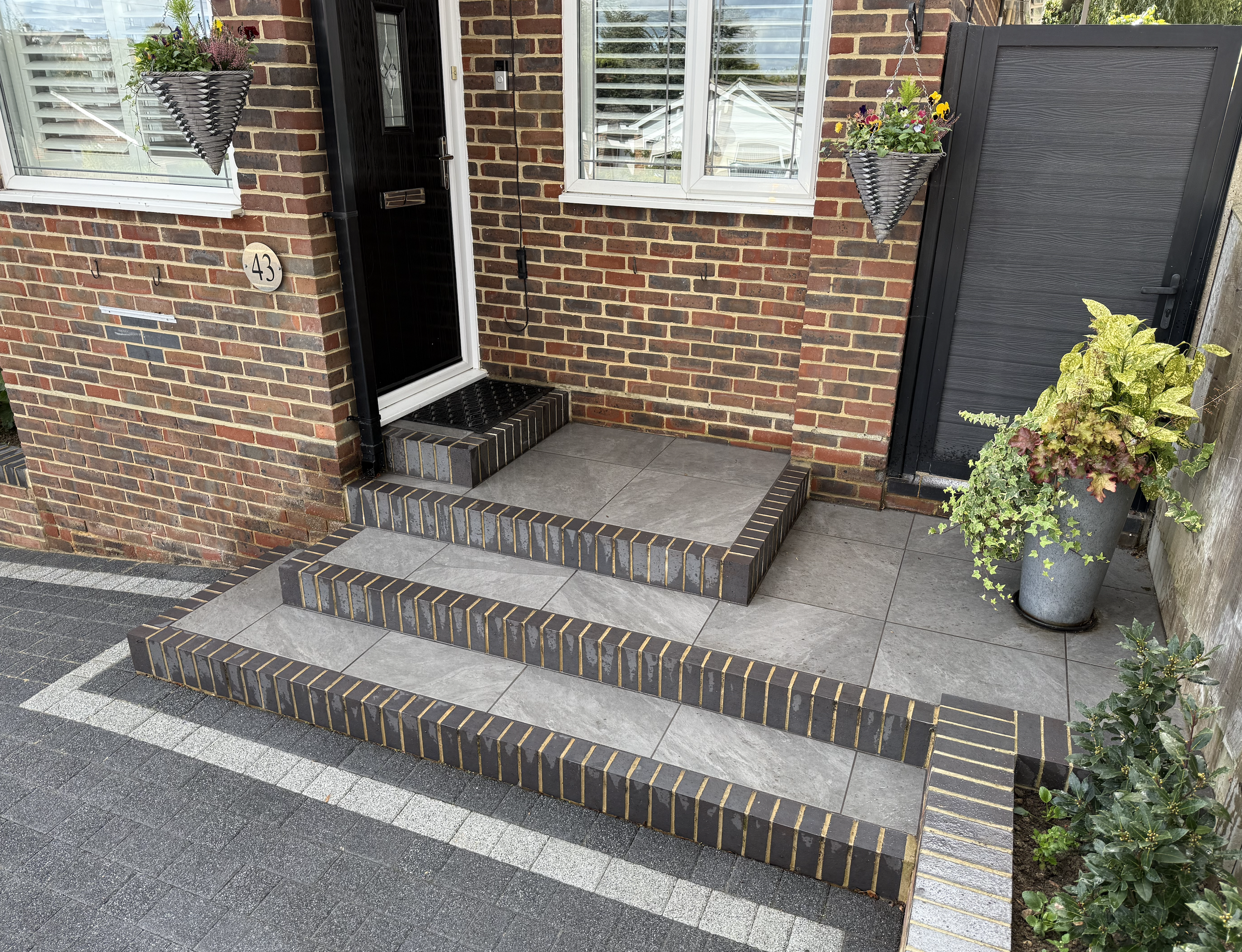 Entrance to a brick house with a small porch, black door, and window screens. The porch has a raised platform with tiled steps bordered with black and yellow bricks. Two hanging flower baskets and a large potted plant are on either side of the door.
