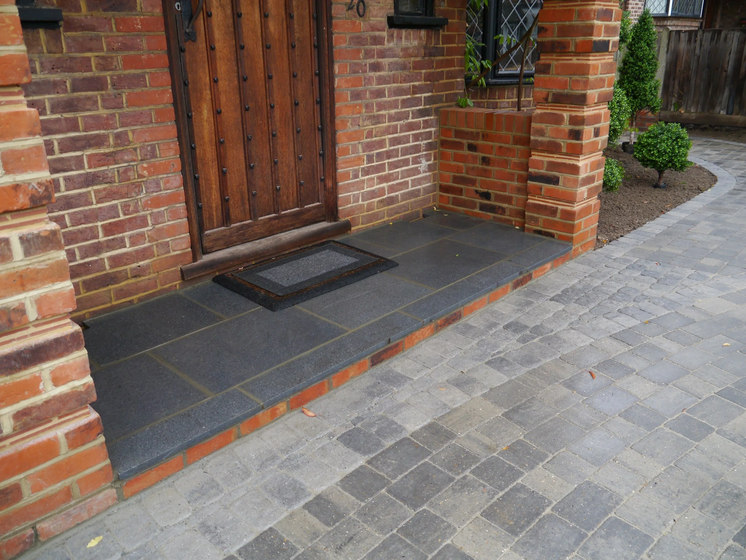 Front porch of a brick house with a wooden door, a small step covered with dark tiles, and a black doormat. There is a small garden with trimmed bushes to the right.