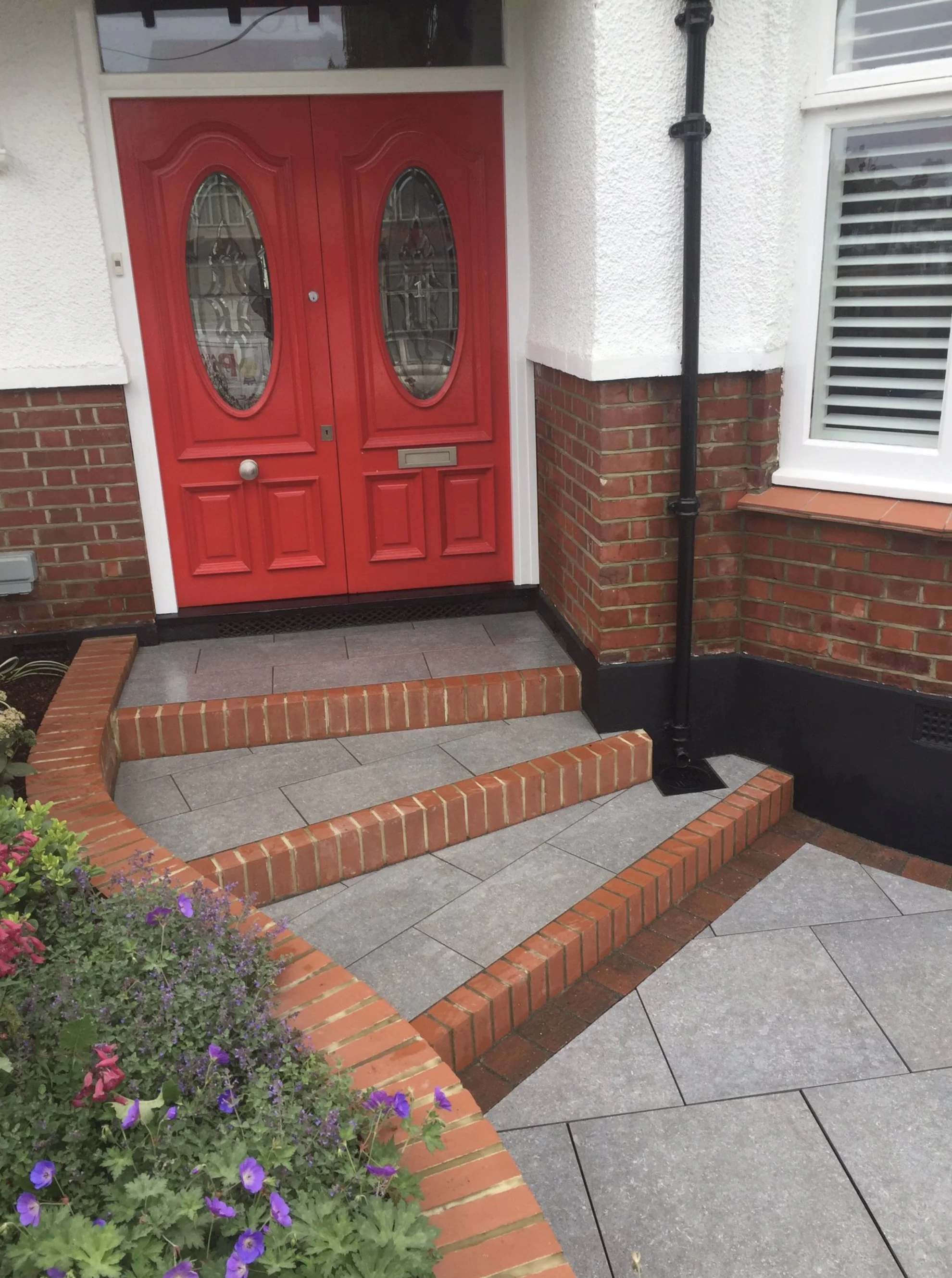 Red front door with oval glass panels, gray tile steps leading up to it, brick and white exterior walls, and a black drainpipe attached to the wall. A flower bed with colorful flowers is in the lower left corner.