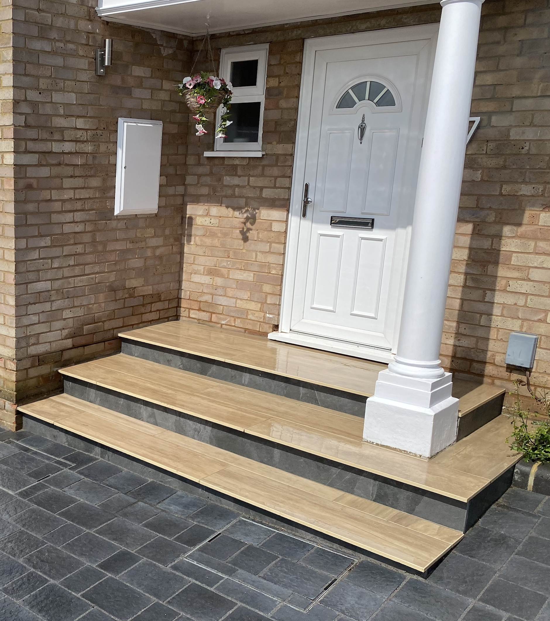 Front entrance of a house with three tiled steps leading to a white front door, a hanging flower basket, a small window, and a brick wall.