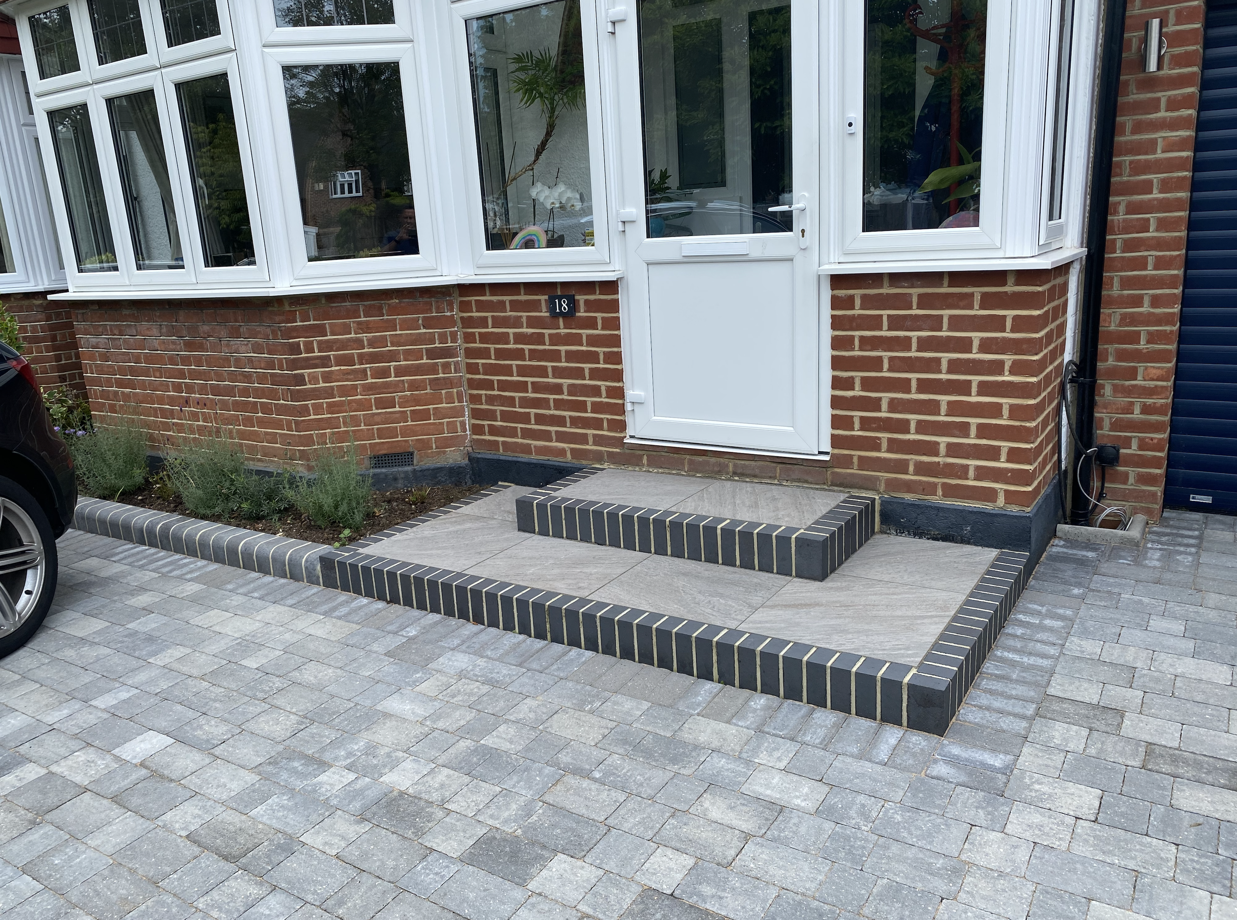 Brick house entrance with a white door and windowed porch, black brick steps and border, stone walkway, parked car, and small garden with plants.