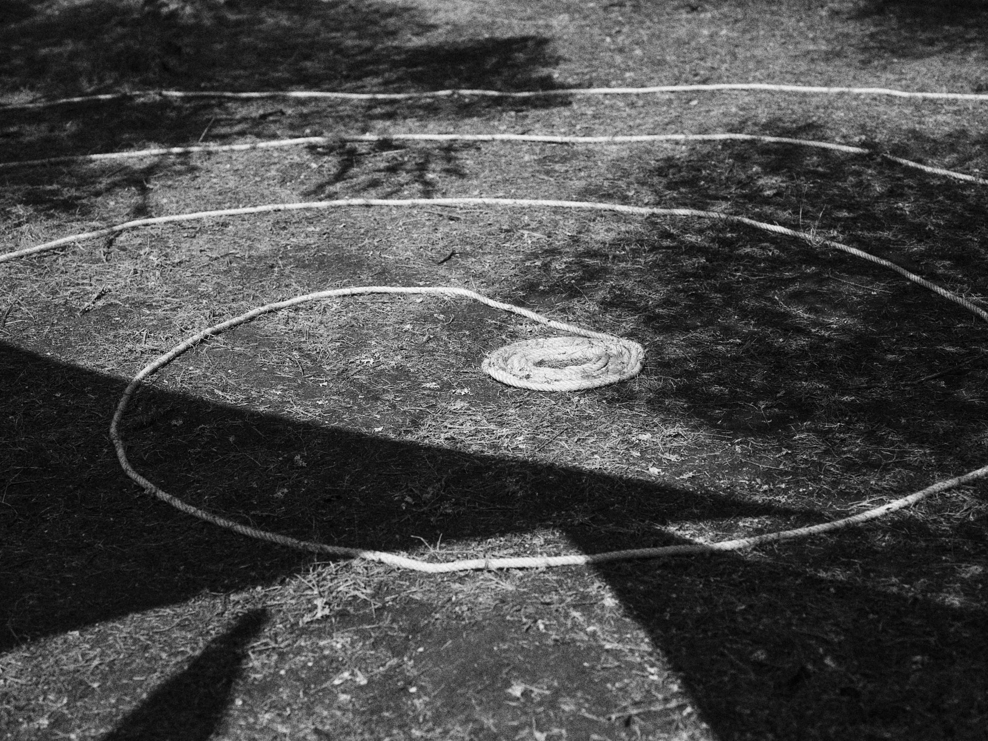 Cuerda en el suelo formando un círculo con una piedra en el centro, sombras y superficie arenosa o de tierra.
