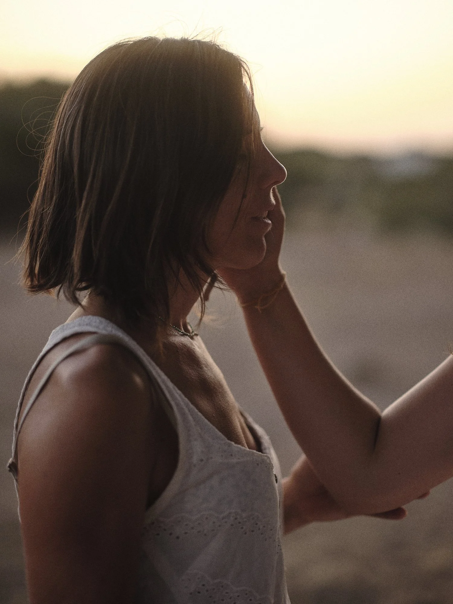 Mujer de cabello castaño oscuro en perfil, con piel clara, usando un top sin mangas blanco, en un entorno al atardecer.