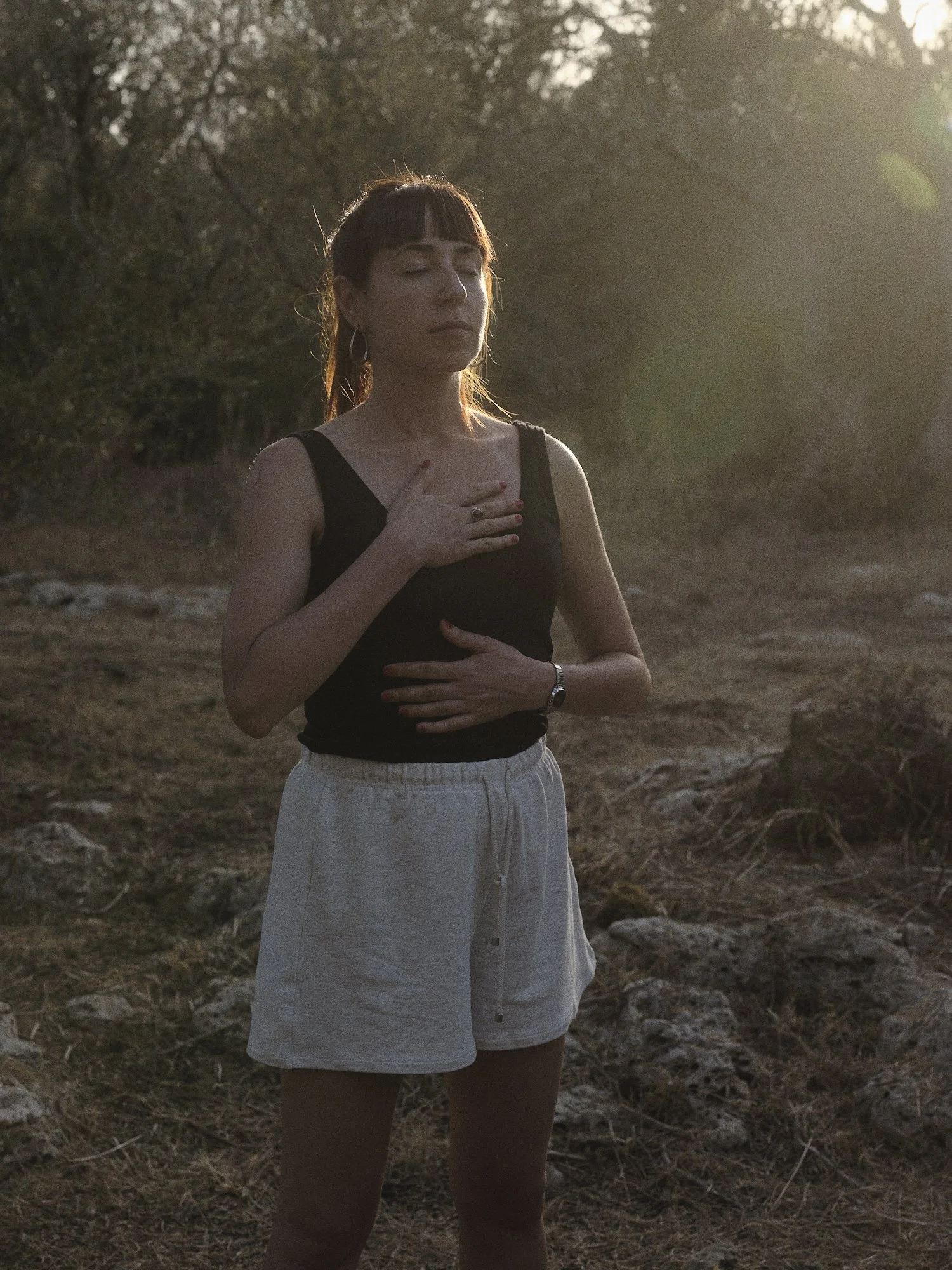Mujer con ojos cerrados, sosteniendo su pecho y abdomen con ambas manos, en un entorno natural al atardecer.