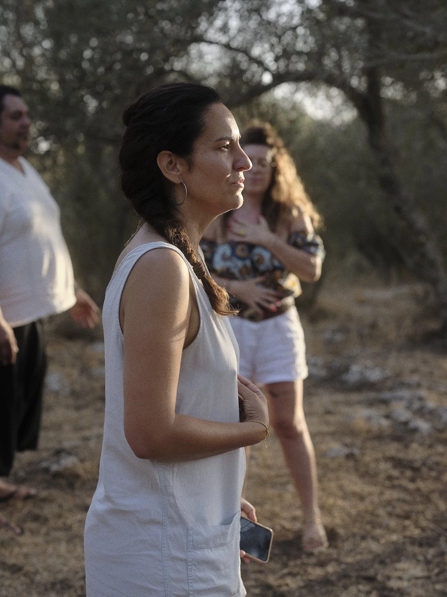 Grupo de personas en un entorno natural, la mujer en primer plano lleva una camiseta blanca, con cabello oscuro peinado en trenza y sostiene un teléfono móvil, mientras otros las observan al fondo.