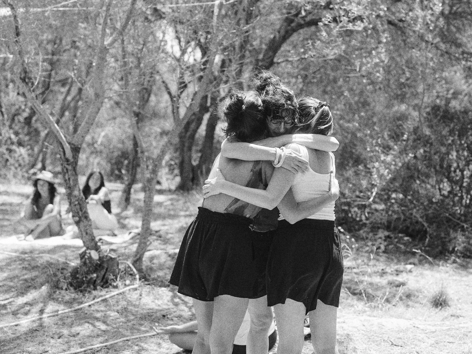 Tres jóvenes mujeres se abrazan en un parque, mientras dos otras mujeres observan desde la distancia, en un entorno al aire libre con árboles y vegetación.