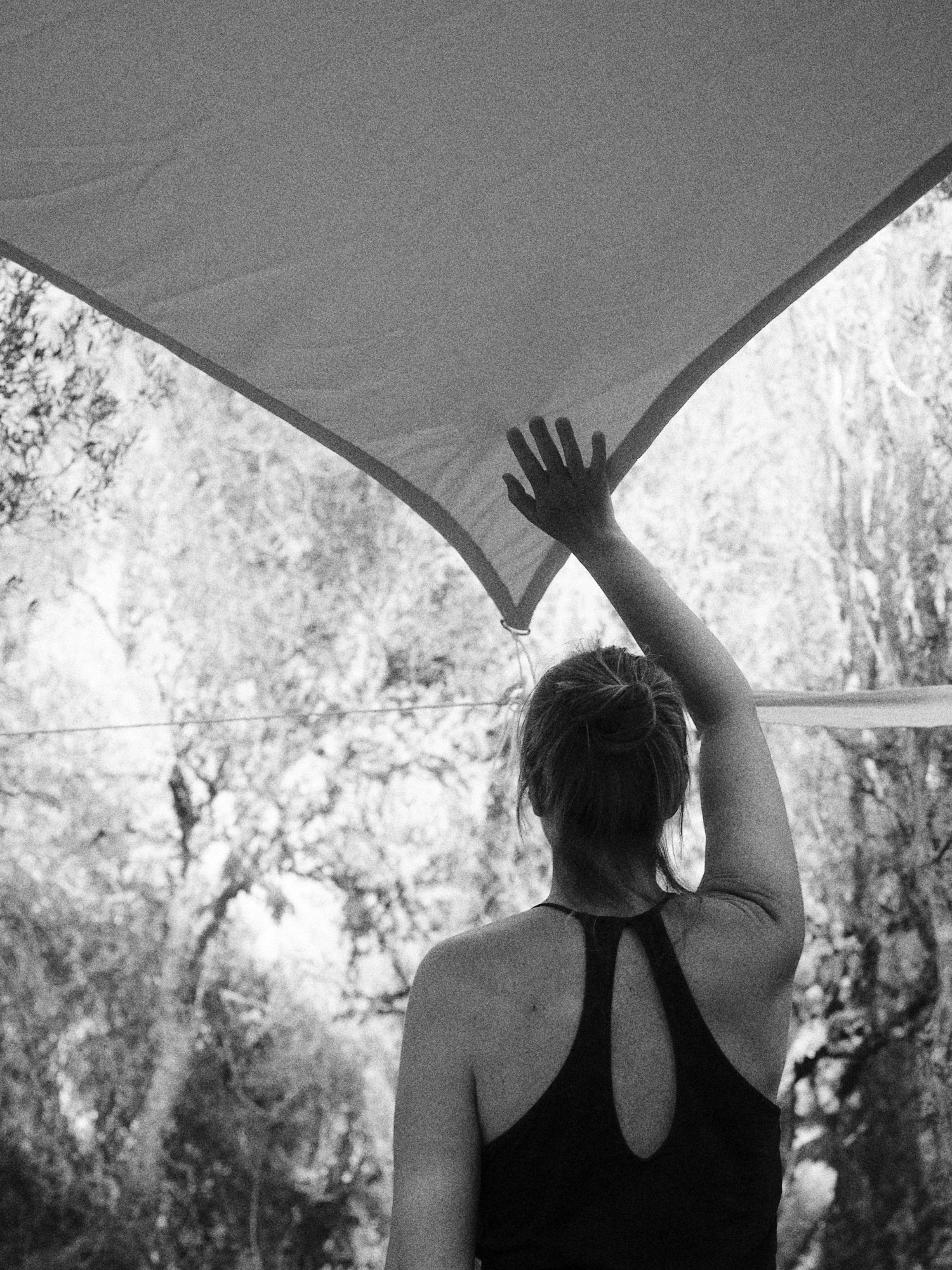 Una mujer vista desde atrás, enganchando una lona o toldo en un espacio exterior con árboles en el fondo, en blanco y negro.