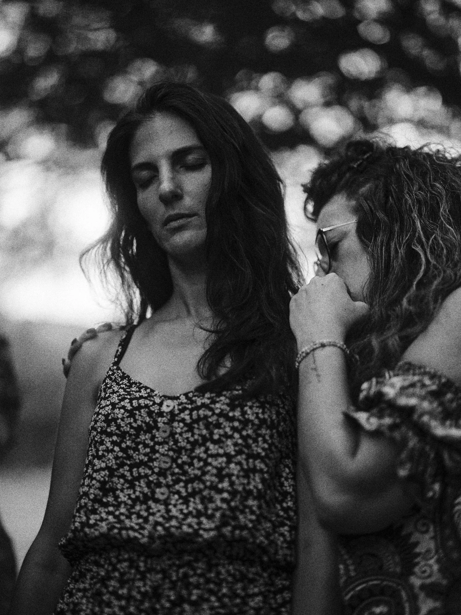 Dos mujeres jóvenes en una conversación emotiva, una de ellas con los ojos cerrados y la otra con gafas, en un entorno al aire libre con árboles y luz de fondo difusa.