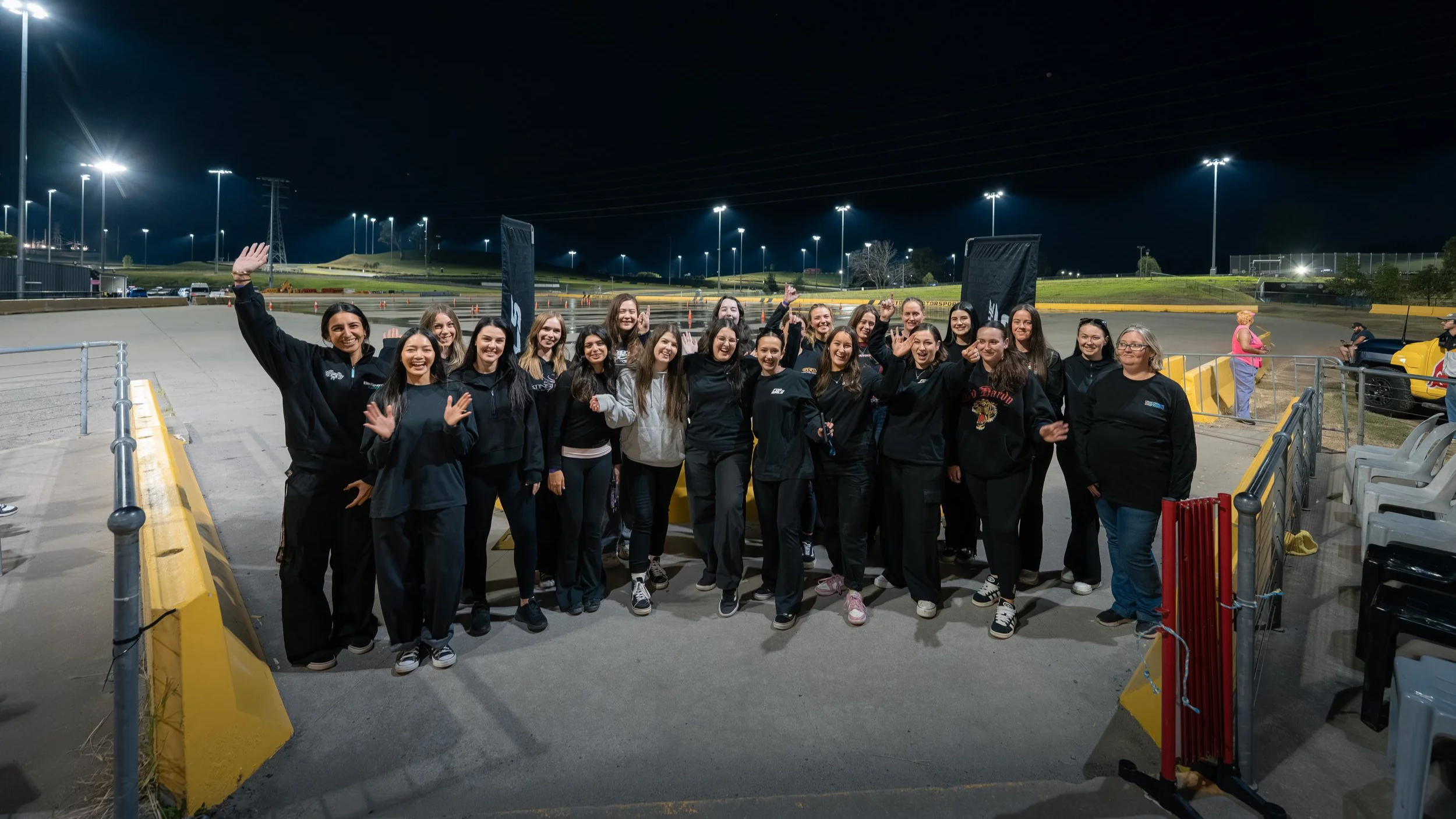 A group of women standing together at night at a racing track, smiling and waving at the camera. The track is illuminated by tall lights, with a few cars and onlookers in the background.