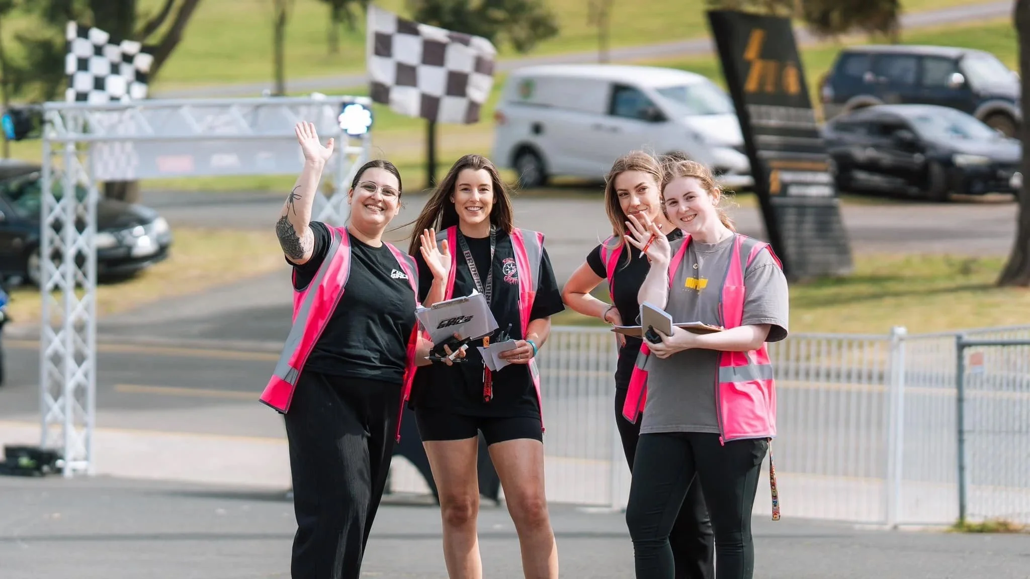 Four women wearing pink vests waving and smiling at an outdoor event, with cars in the background.