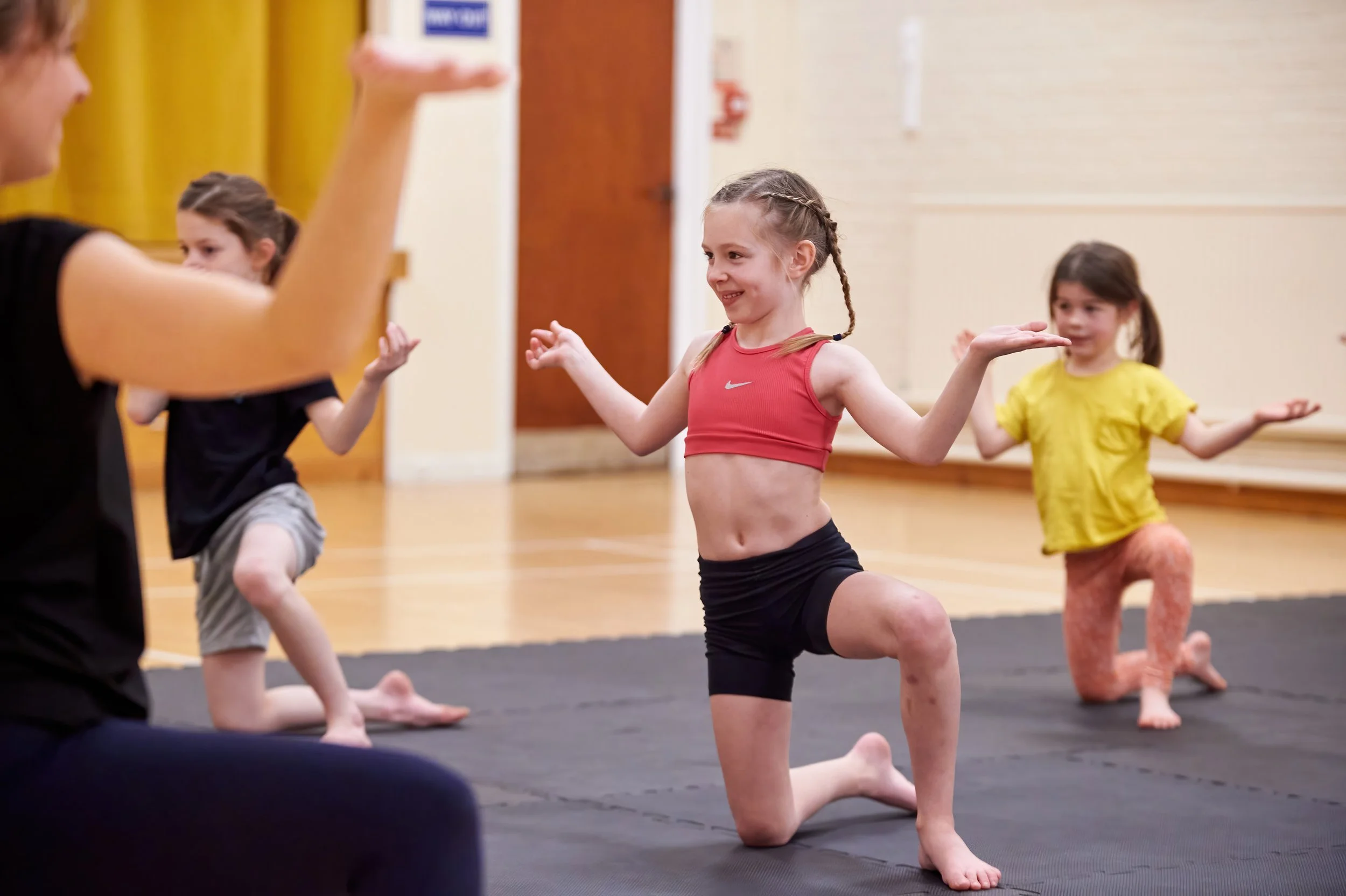 Young girls participating in a yoga class, kneeling on a black mat, smiling, and practicing poses in a gymnasium with wooden floors and yellow curtains.