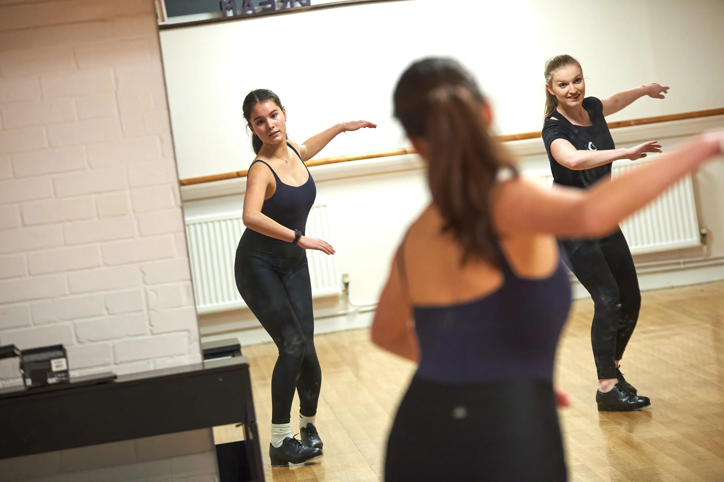 Two women with brown hair in a dance studio, both balancing on one leg with arms extended for ballet practice, as another woman with brown hair in a ponytail observes in front of a mirror.