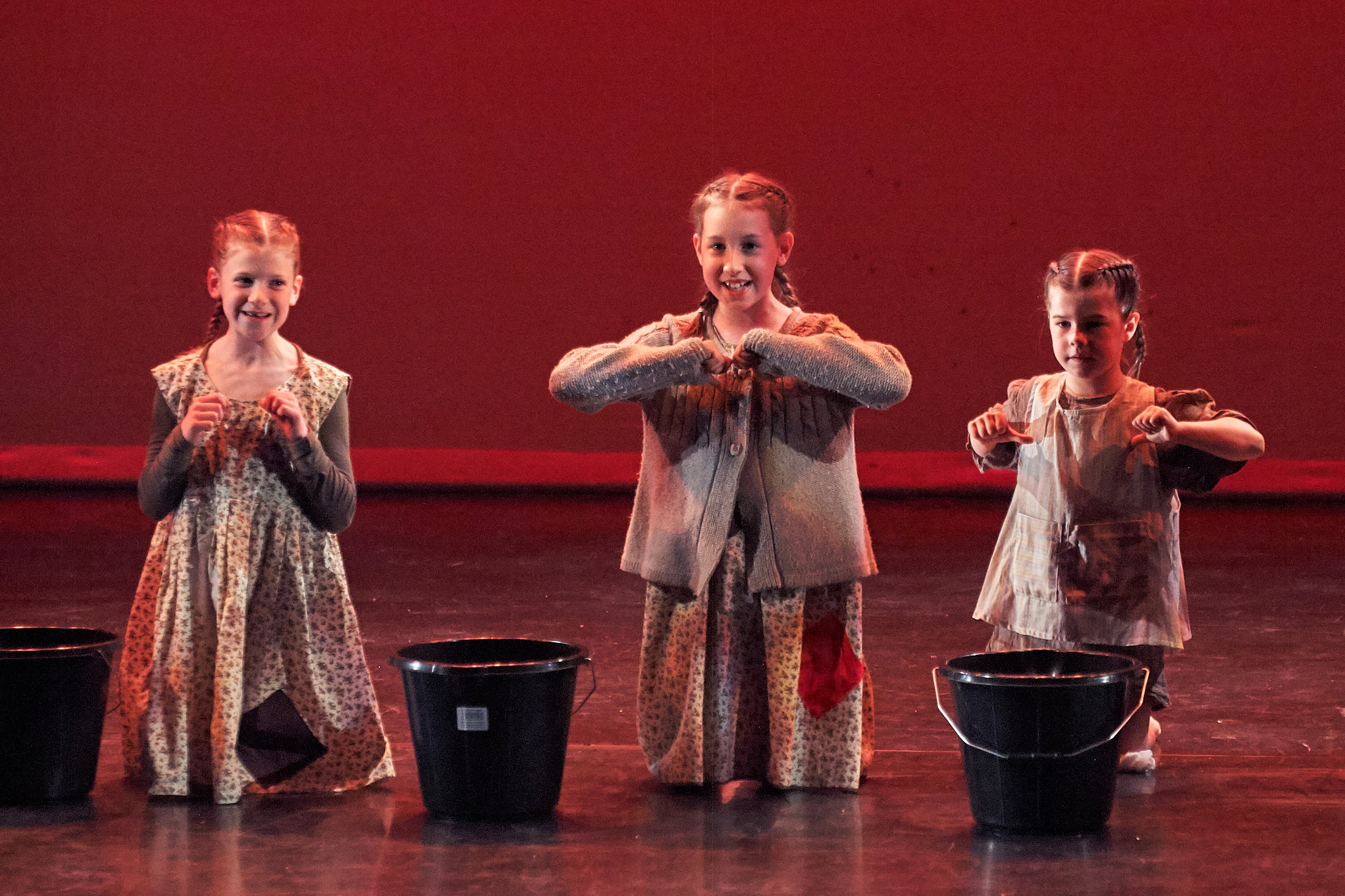 Three young girls in vintage-style dresses standing on stage, smiling, with buckets in front of them, illuminated by stage lights against a dark background.