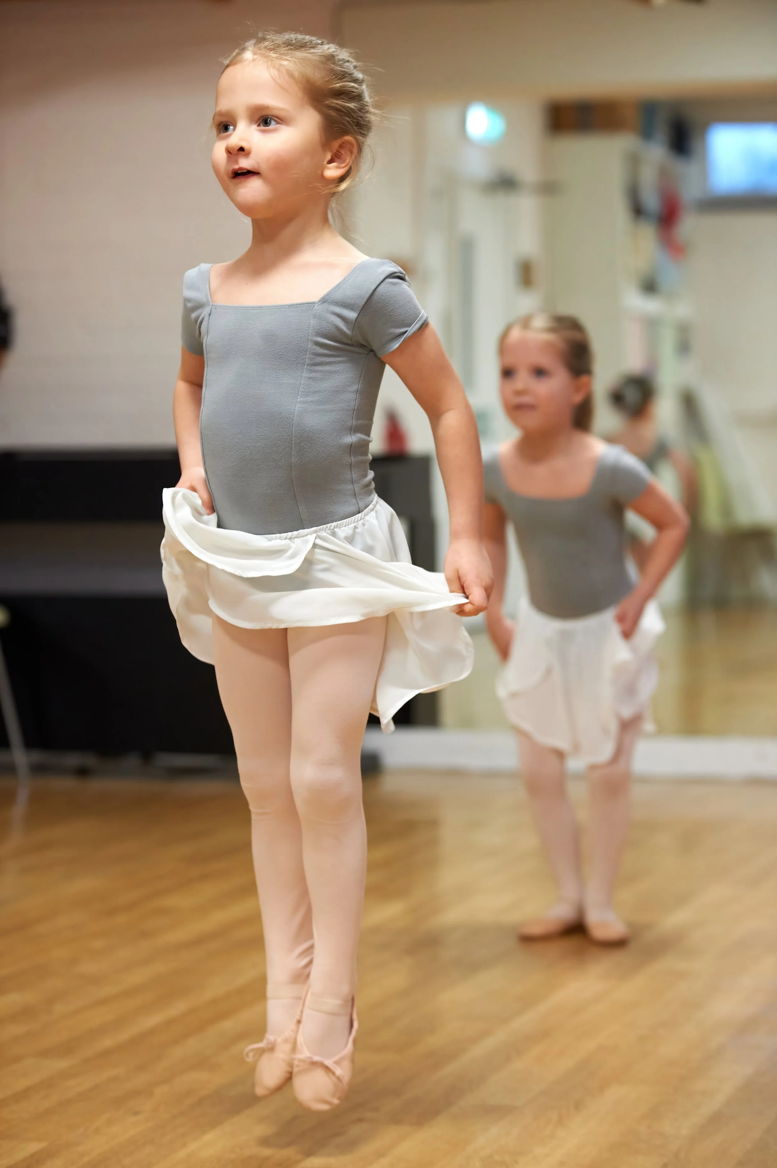 Young girls in ballet costumes practicing ballet in a dance studio with wooden floors.