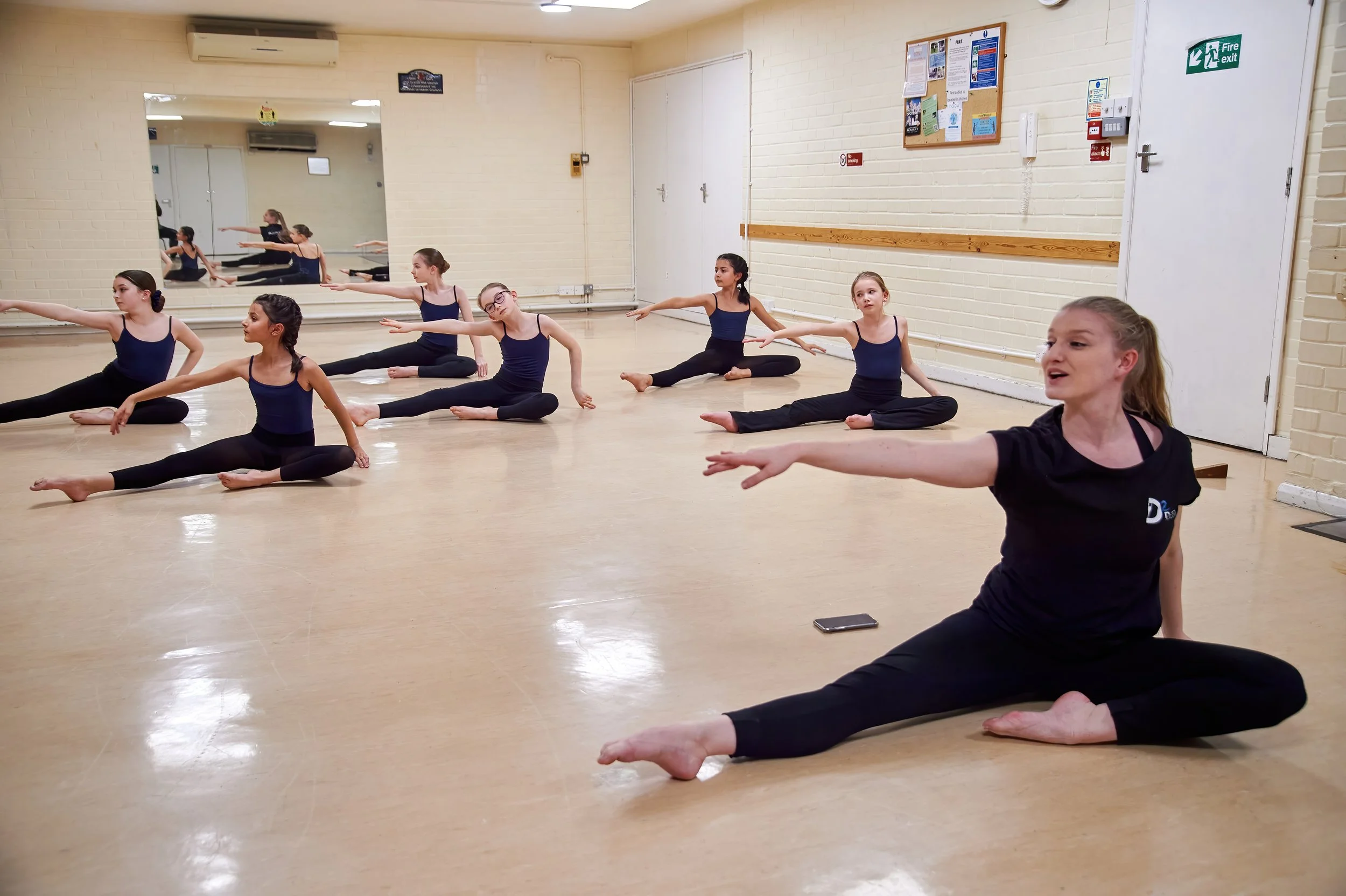 Young girls practicing ballet dance in a dance studio with a mirror on the wall, wearing navy blue leotards and black leggings.