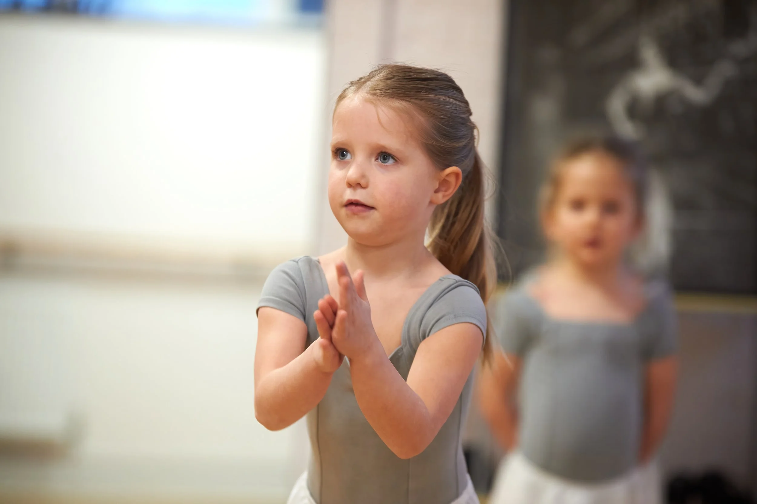 Young girl practicing ballet, wearing gray leotard, standing with hands together in a dance studio with another girl blurred in the background.