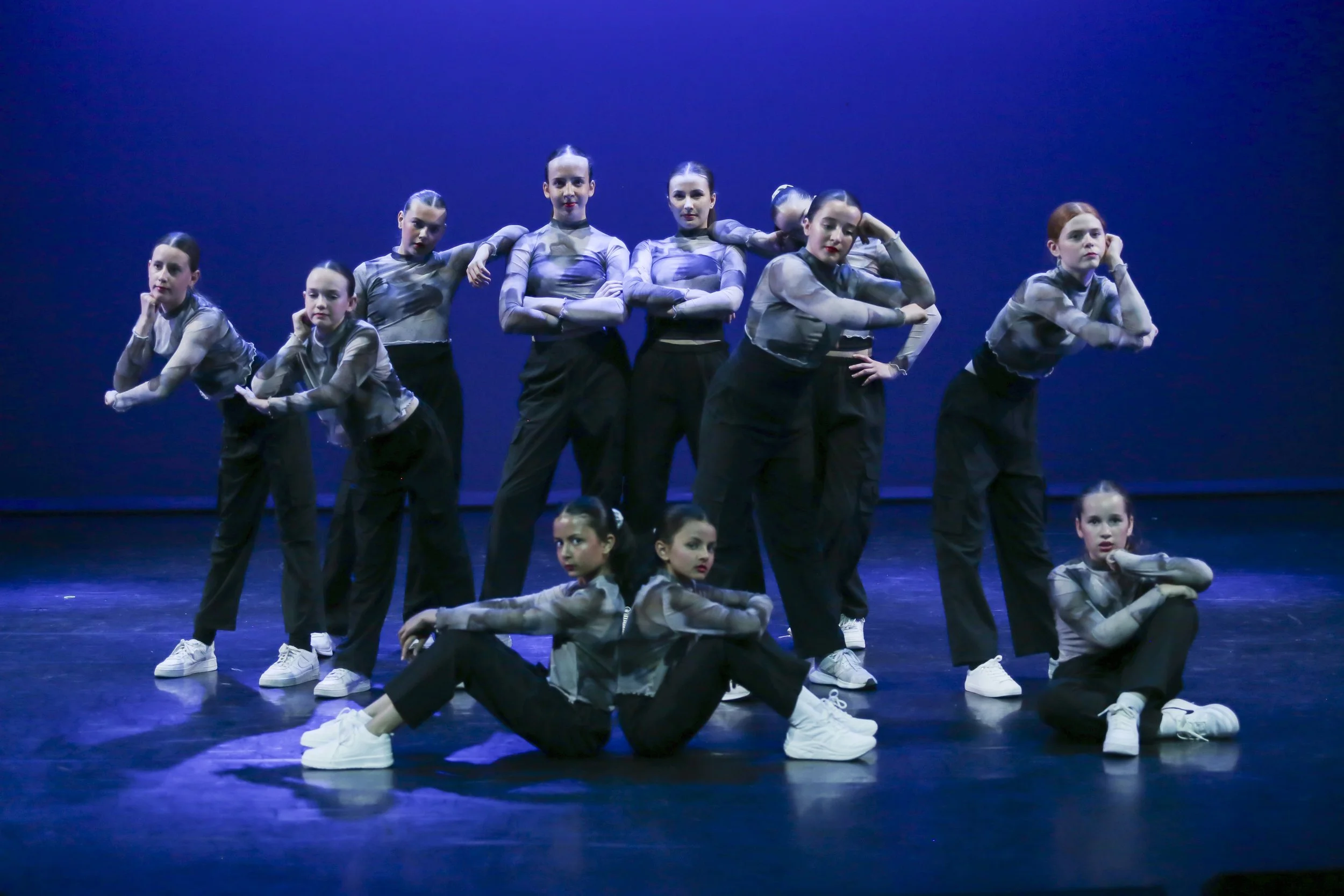 Group of young female dancers in matching costume poses on stage with a blue background.