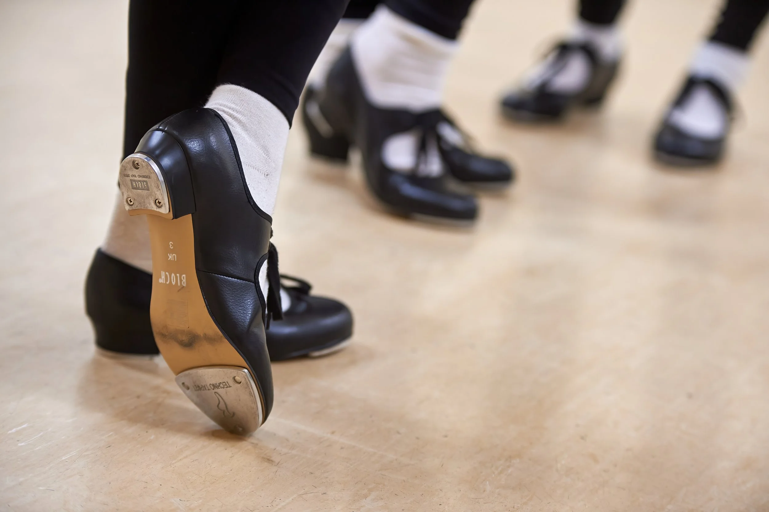 Close-up of a prosthetic leg with a black and beige color scheme, showing a plastic foot touching the floor. In the background, multiple people are standing on ice skates, with blurred ice-skating rink surroundings.