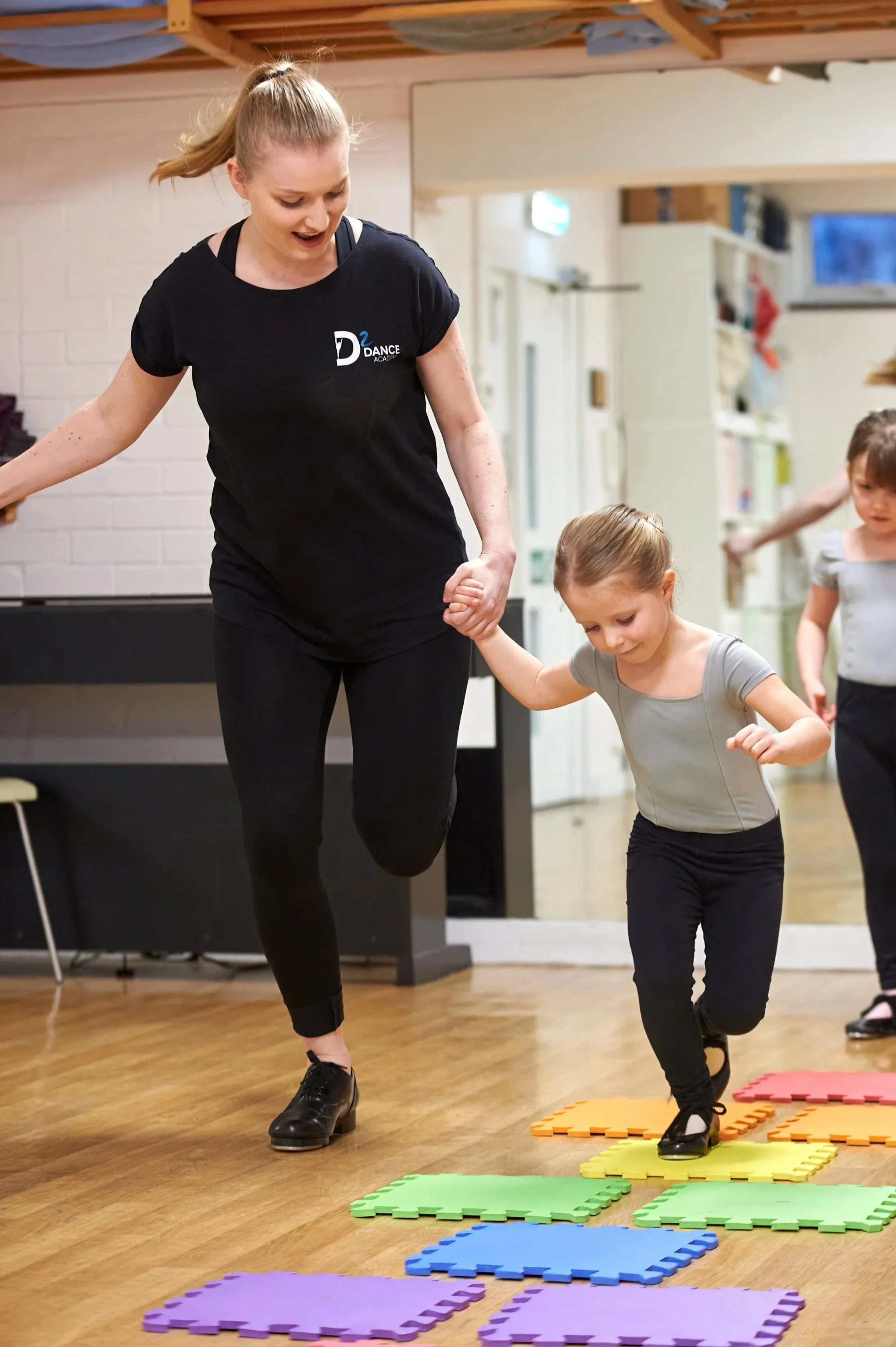Dance instructor guiding young girl through dance moves in a dance studio with colorful foam mats on the wooden floor.