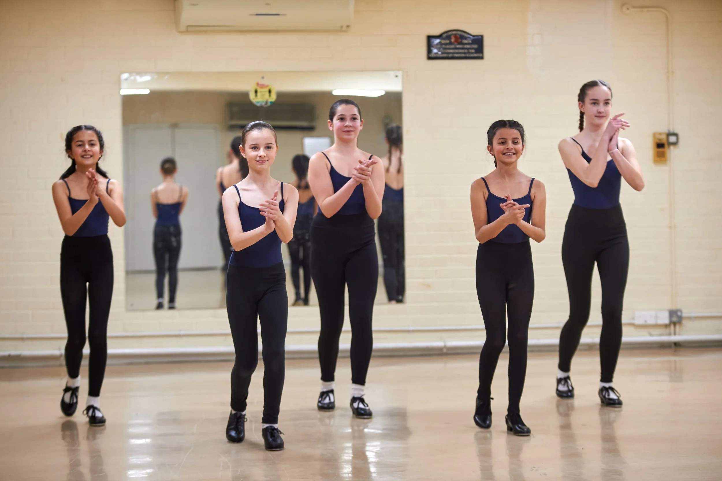 Group of five young ballet dancers standing in a dance studio, clapping, dressed in navy blue leotards, black leggings, and ballet shoes. Mirror behind them reflects their back view.