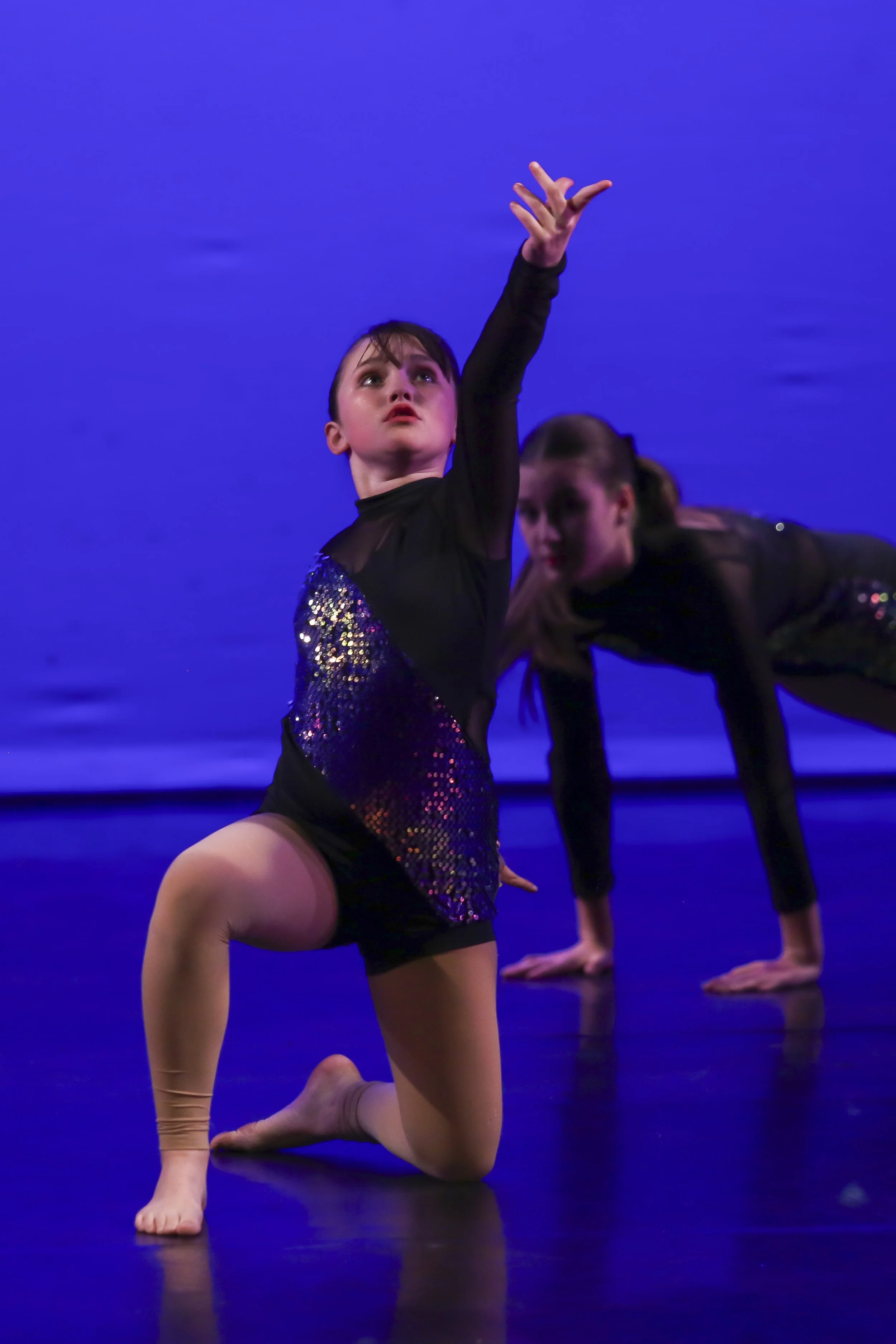 A young girl in a black and purple sparkly dance costume performs a dance move on stage with a blue background, kneeling with one bent knee and one hand raised.