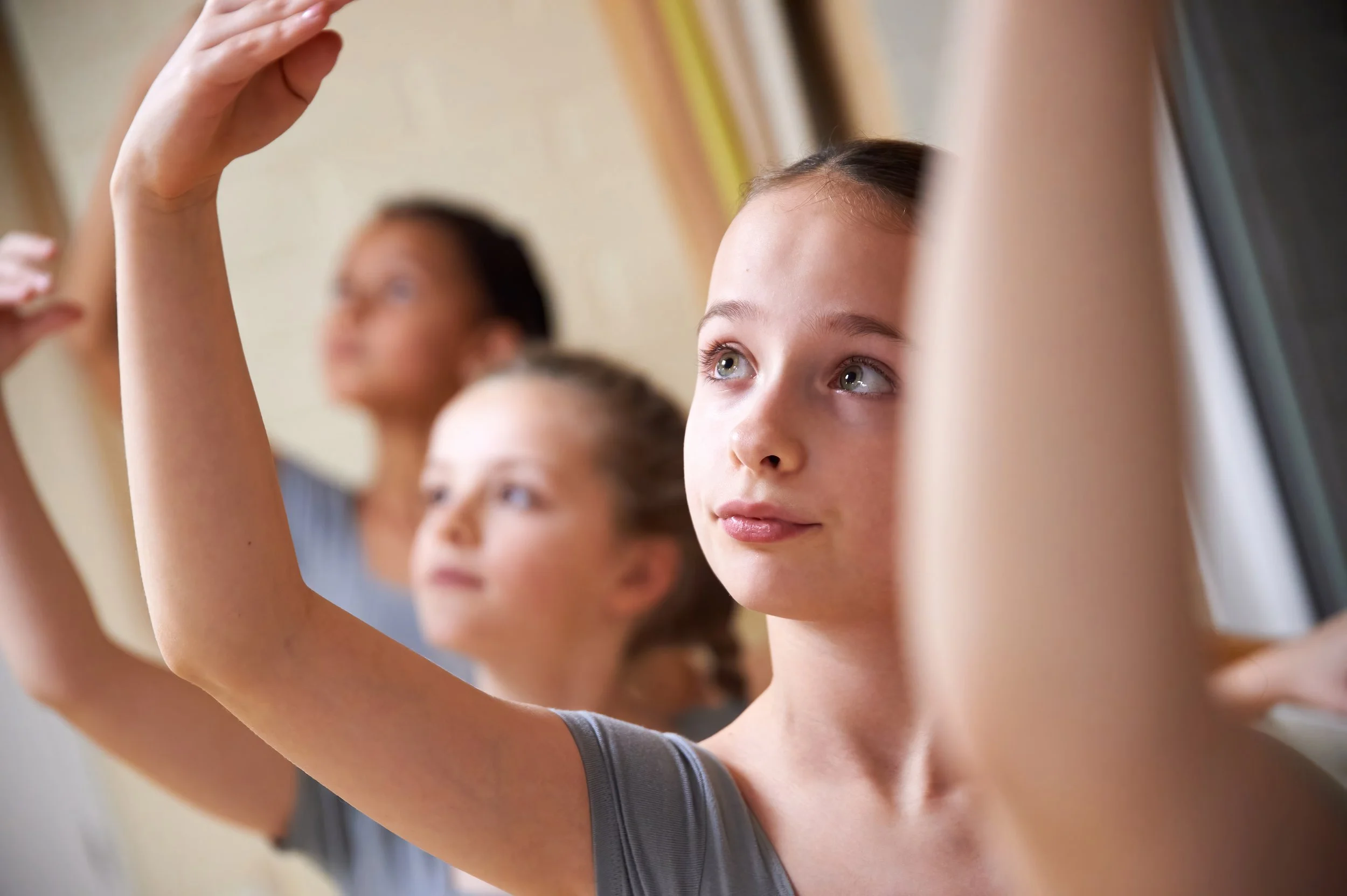 Young girl with brown hair and hazel eyes raising her hand in a classroom with other students in the background.