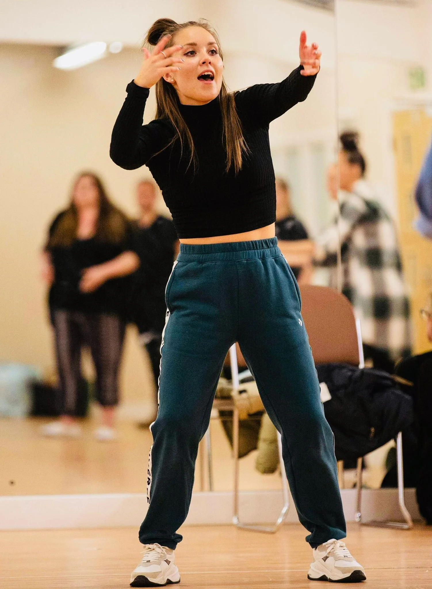 Young woman in black long sleeve top and blue pants dancing or performing in a room with wood flooring, with blurred group of people watching in the background.