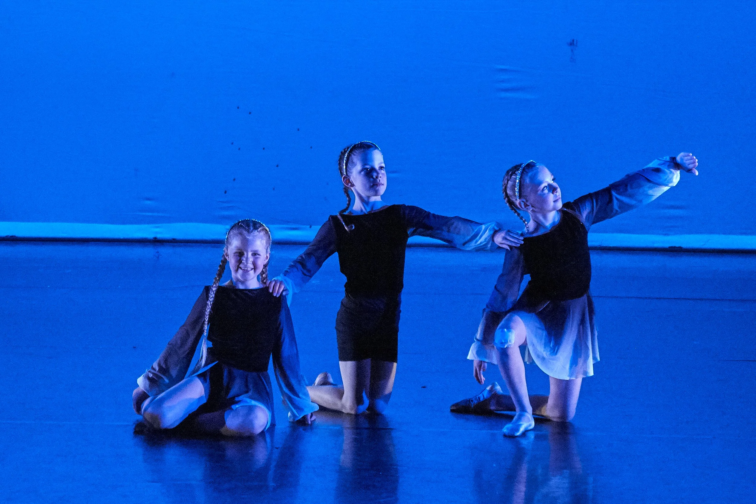 Three young girls in black and white costumes performing a ballet on stage with blue lighting.