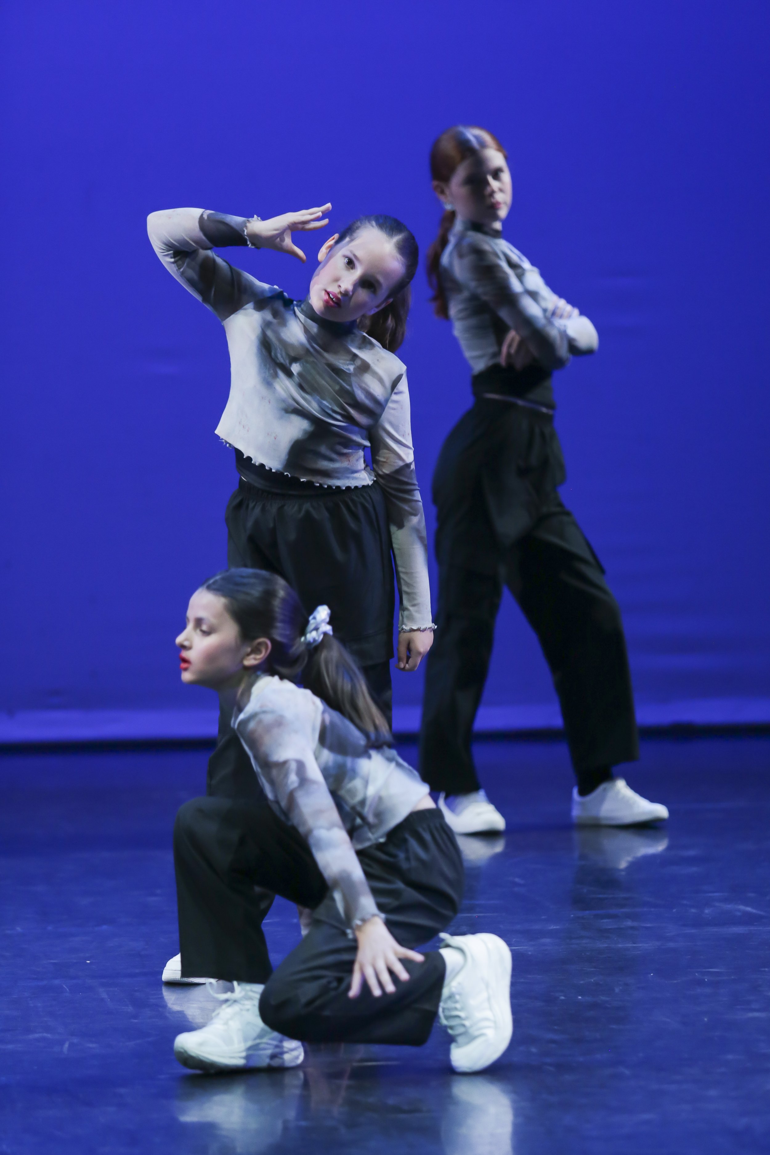 Four young girls in matching outfits performing a dance routine on a stage with a blue background. Two girls are standing with their arms crossed and one girl is kneeling on the floor with her hand on her knee, while another girl is bowing down.