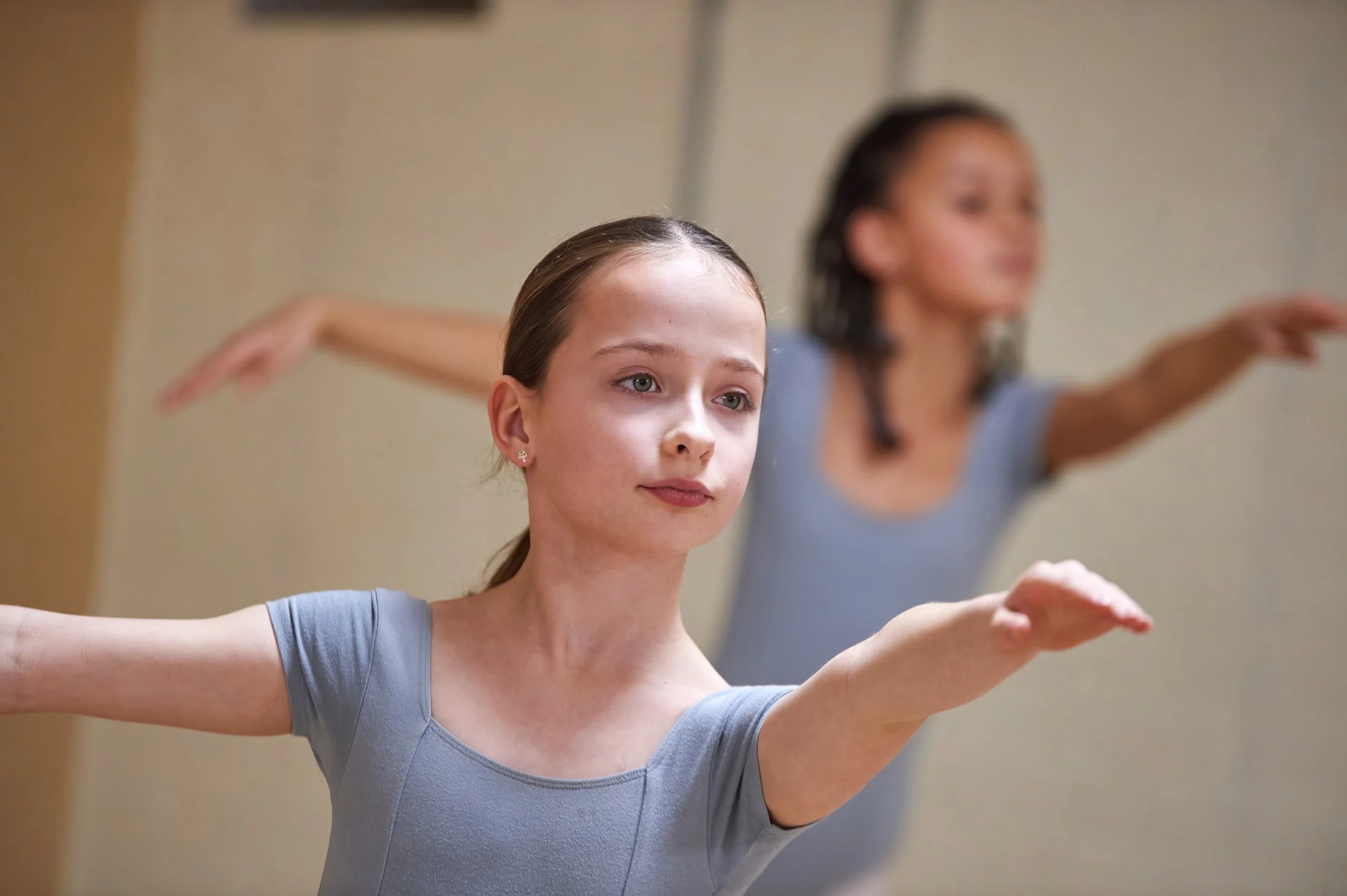 A young woman with light brown hair in a bun practicing ballet in a dance studio, wearing a gray leotard with arms extended outward.