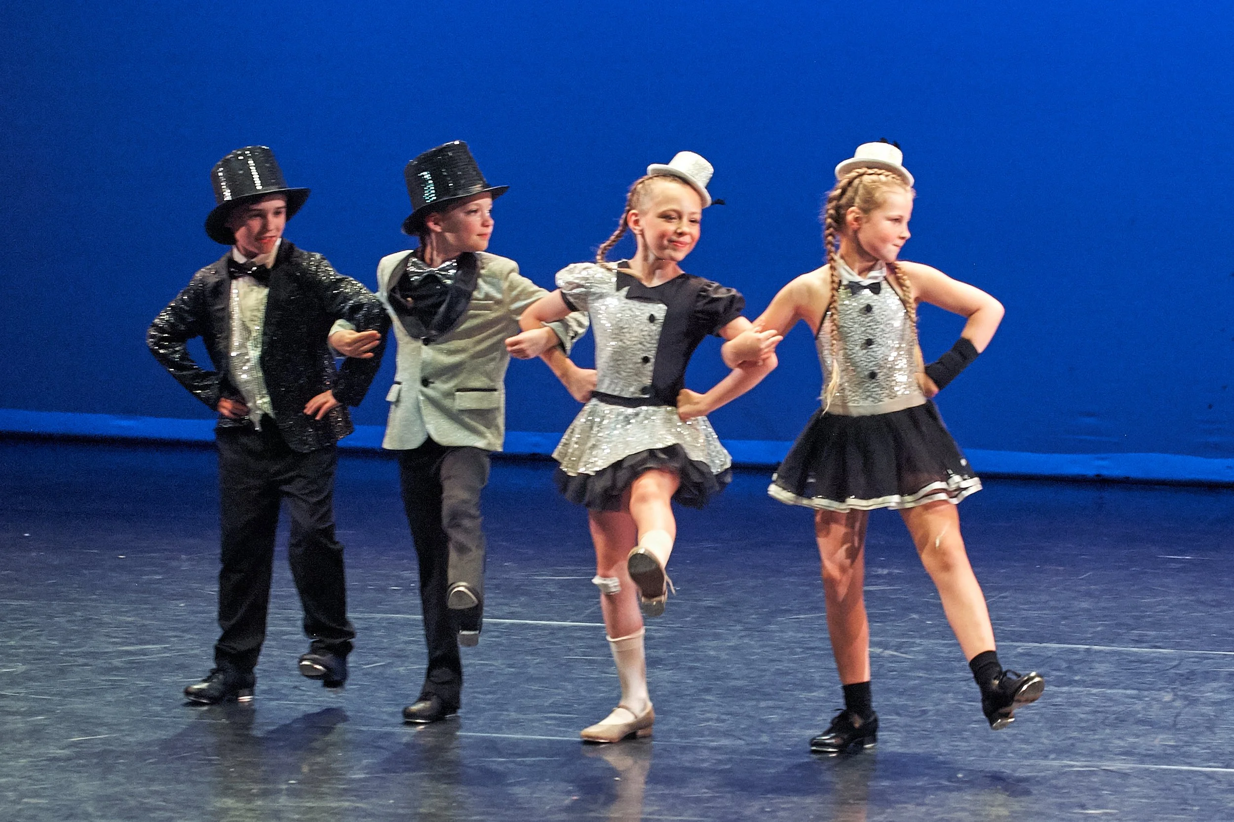 Young children performing a choreographed dance on stage, dressed in costumes and top hat accessories, with a blue background.