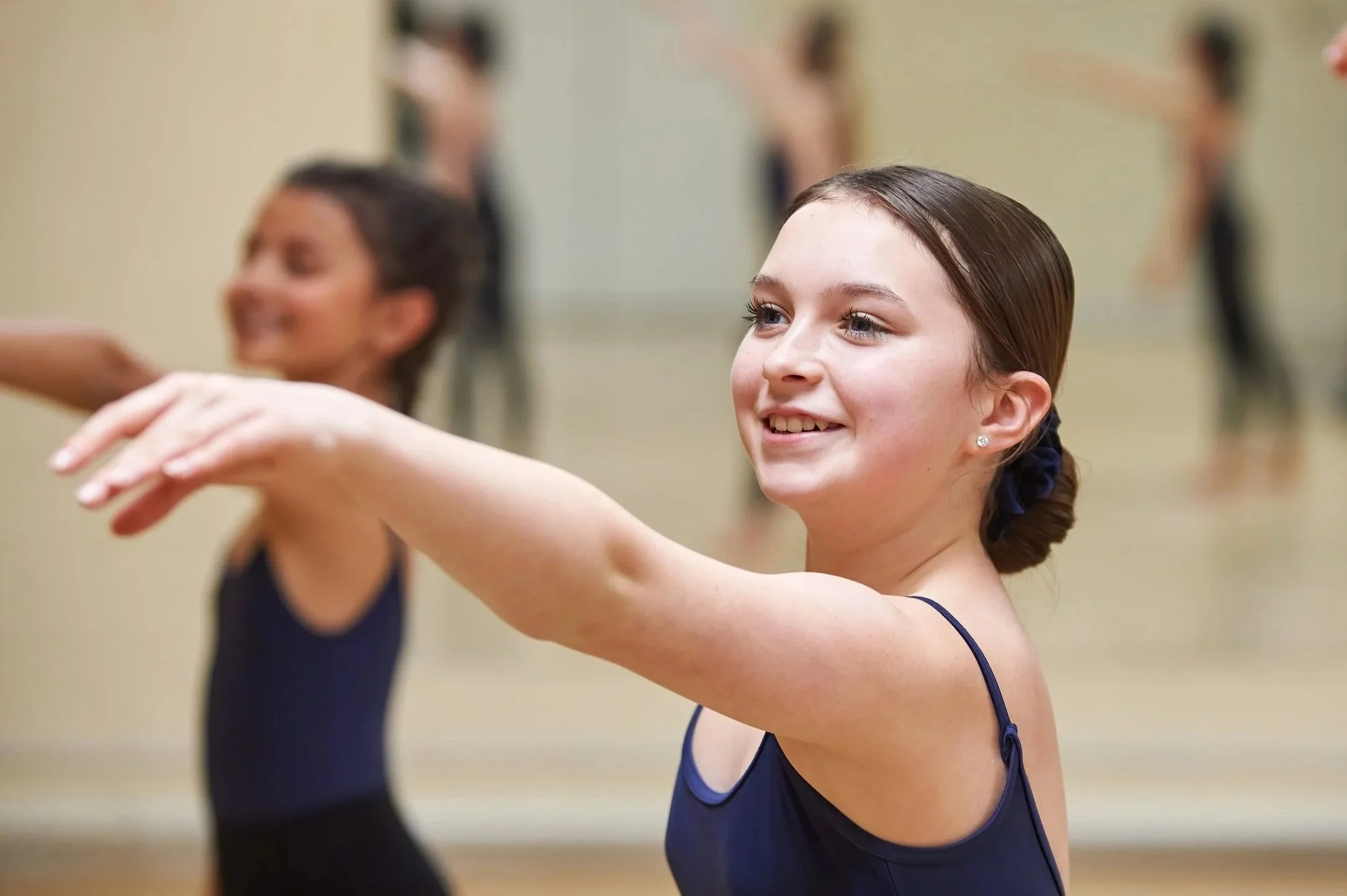 Two young women in ballet class, with the woman in the foreground smiling and the woman in the background out of focus.