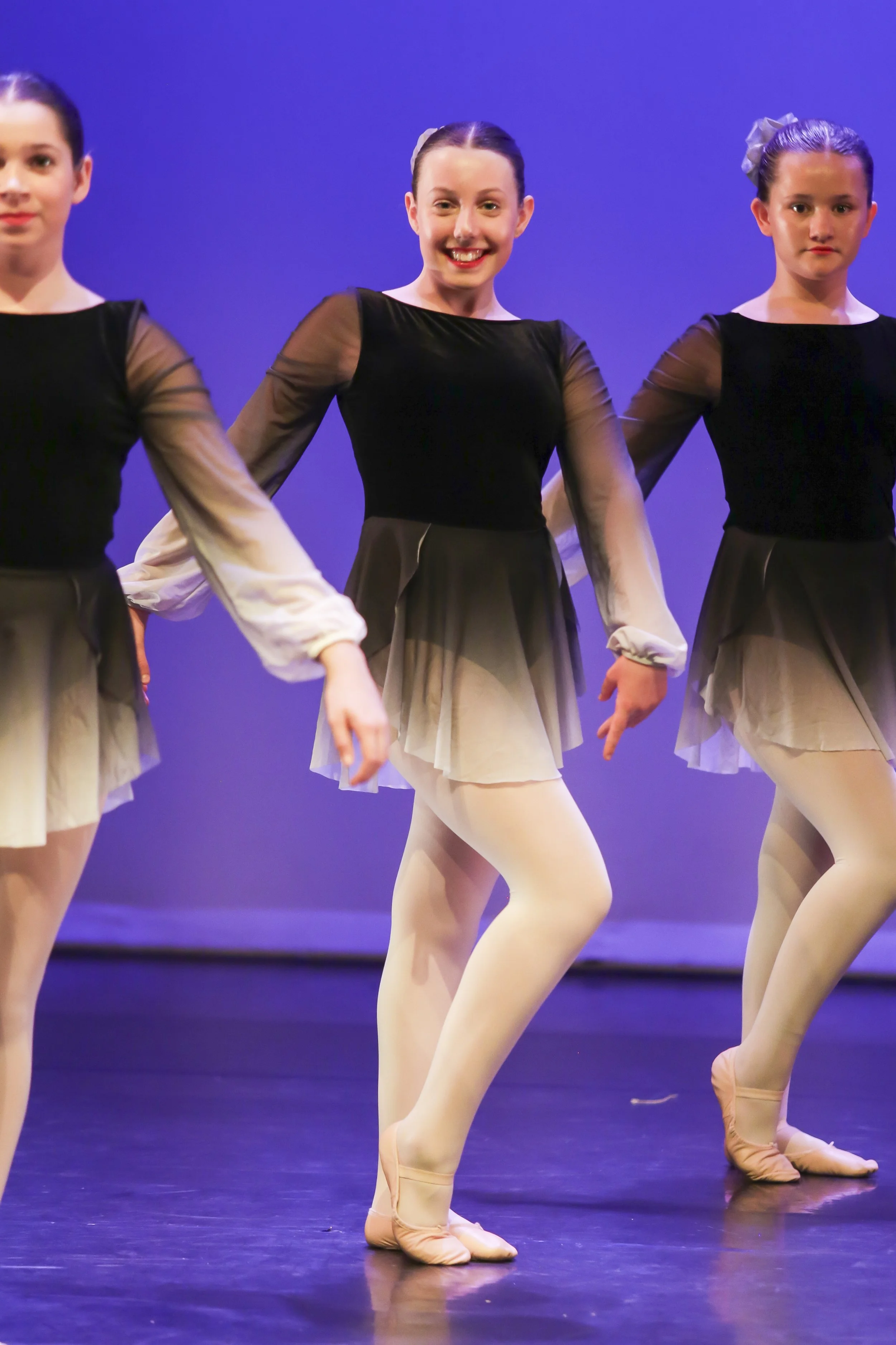 Ballet dancers performing on stage, wearing black and beige costumes with sheer long sleeves and ballet slippers, against a blue background.