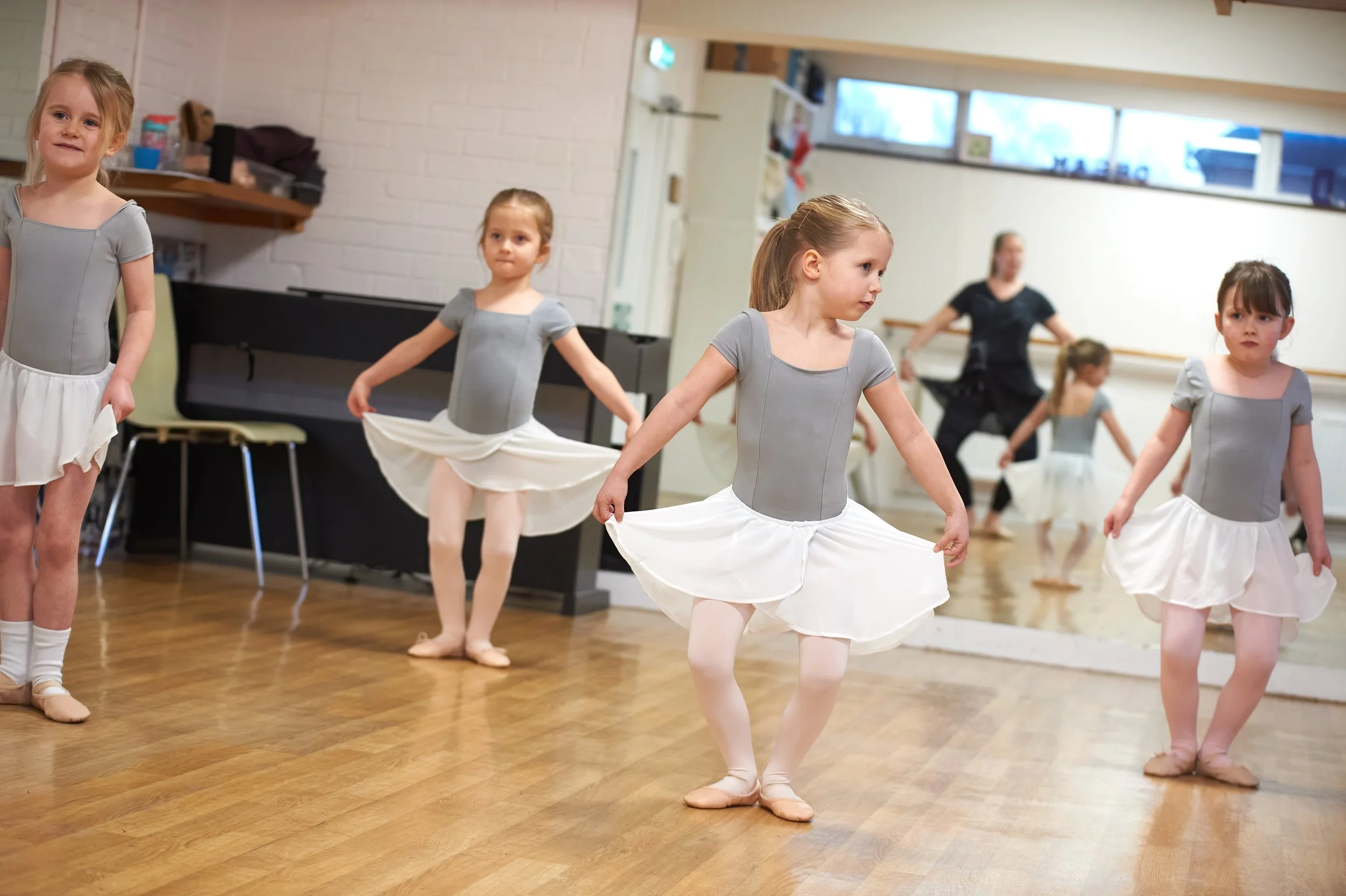 Children in ballet costumes practicing in a dance studio, some holding skirts, with an instructor observing in the background.