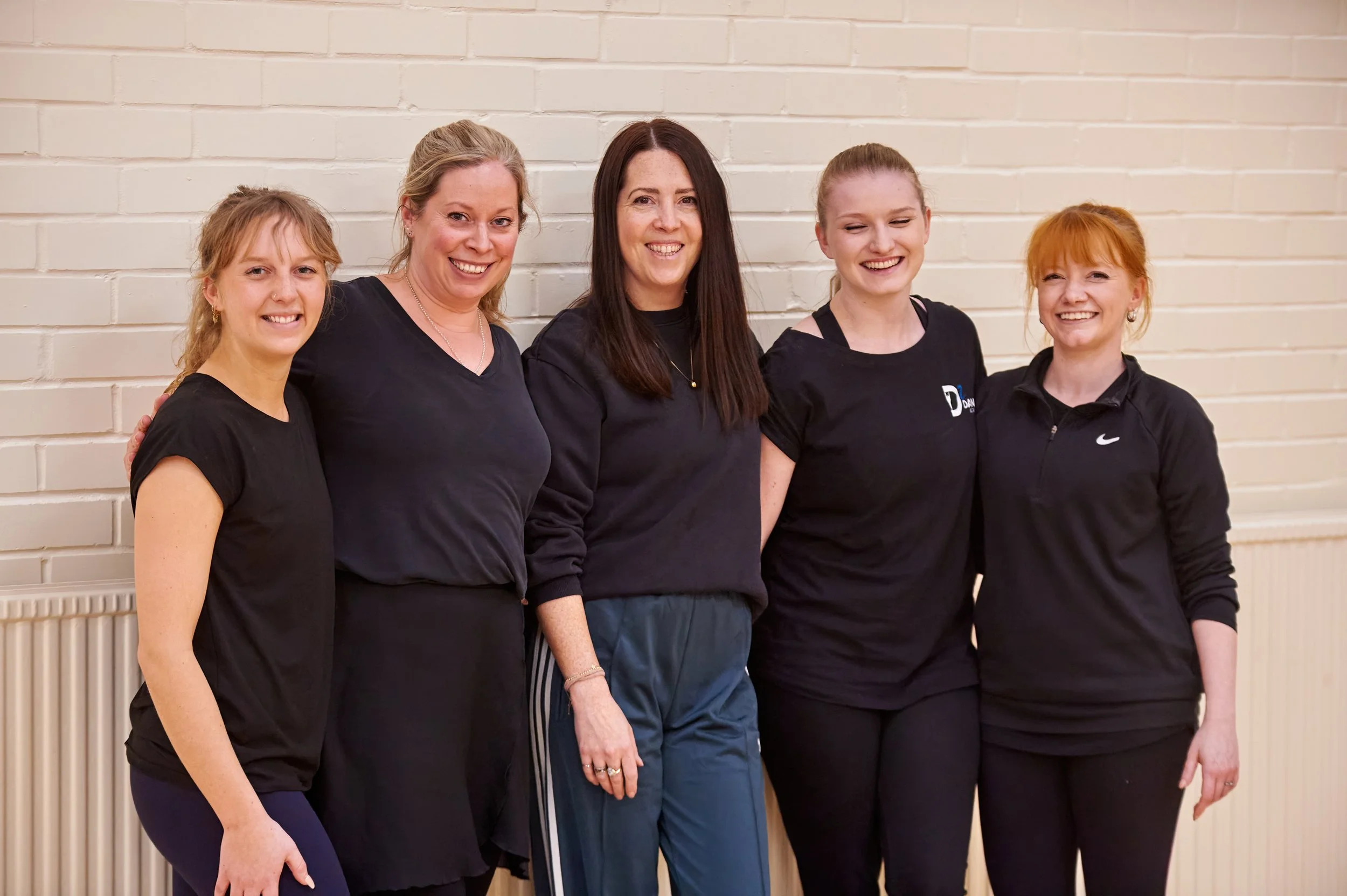 Group of five women standing close together against a white brick wall, smiling at the camera.