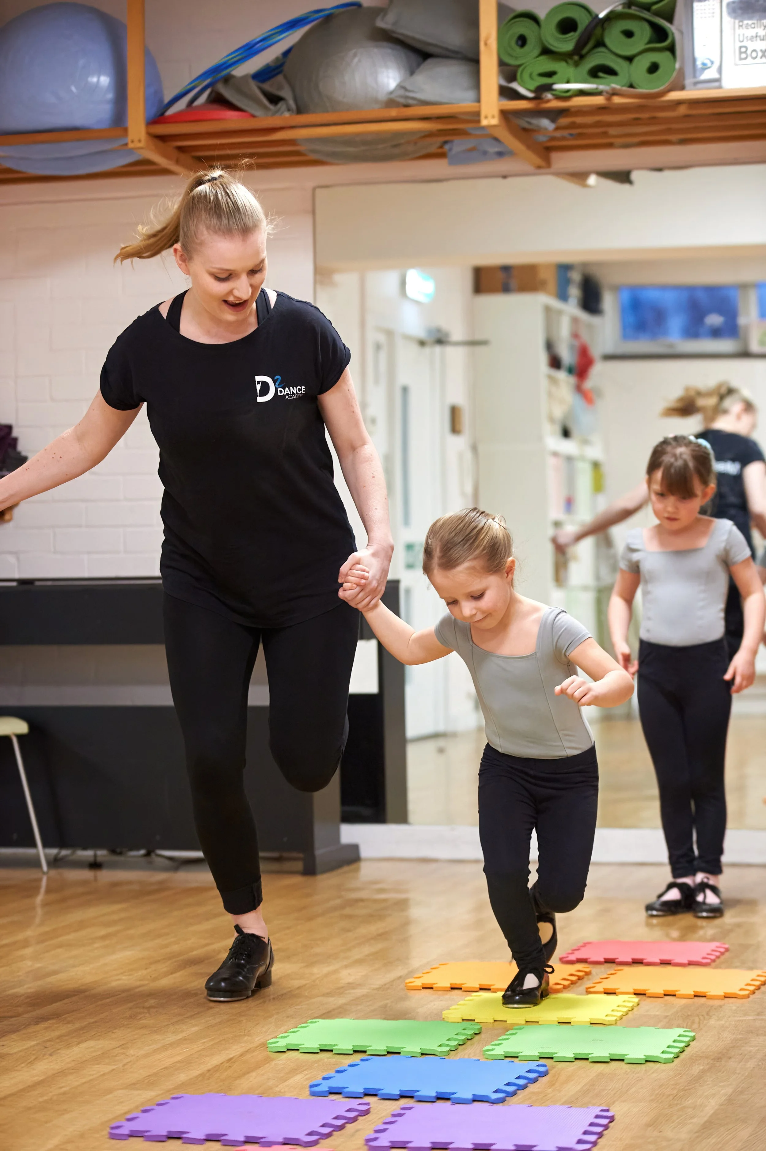 A dance instructor and children practicing dance moves on colorful foam mats in a dance studio. The instructor is holding a child's hand as they balance on one foot with raised knees, with other children waiting their turn.