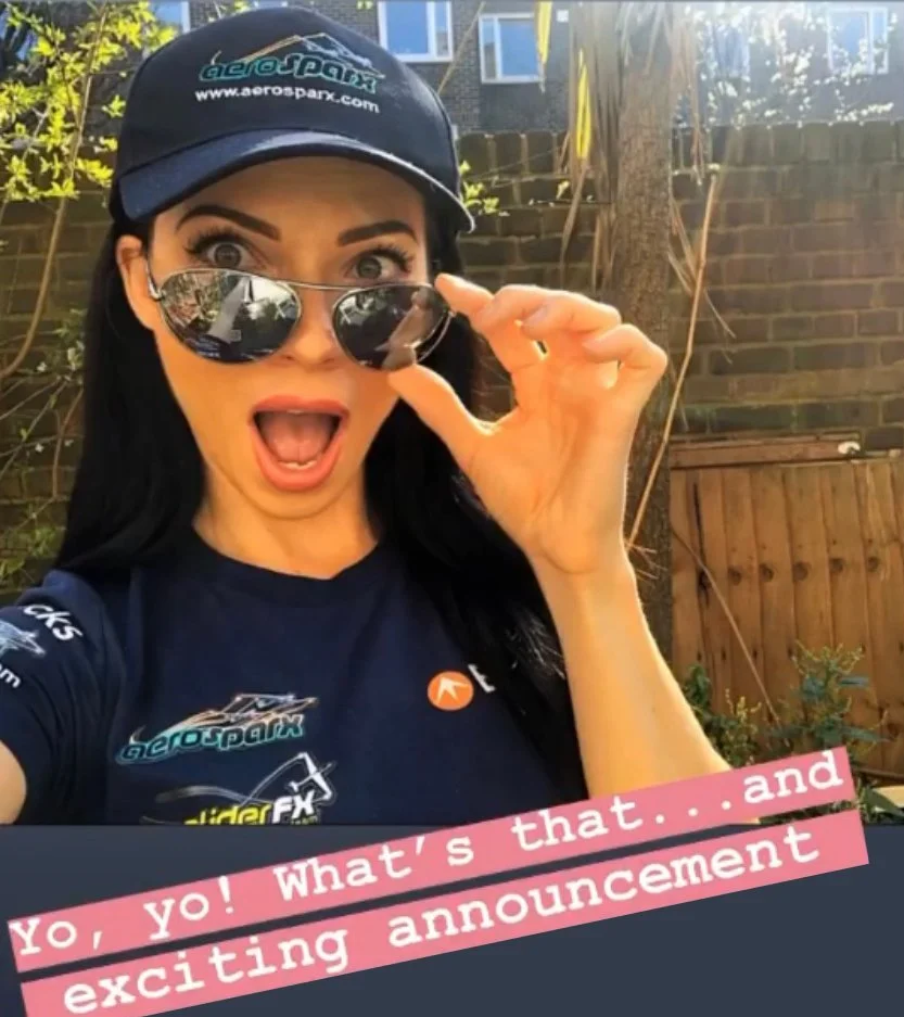 A woman with long dark hair, wearing a black cap and reflective sunglasses, is holding her sunglasses with an excited expression. She is outdoors, with a brick wall, a tree, and a wooden fence in the background. She is wearing a navy blue t-shirt with various logos and a pink overlay caption that says, "Yo, yo! What’s that. . . and exciting announcement."