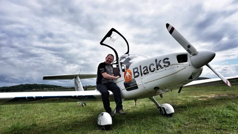 A man sitting on the wing of a small white airplane with an open cockpit door, on grassy ground under a cloudy sky.