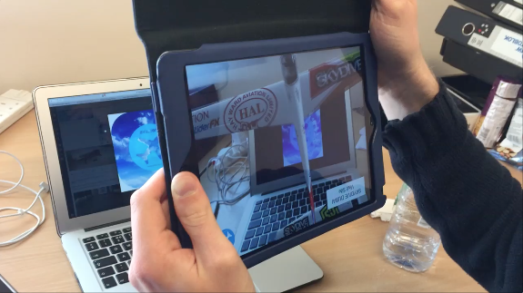 Person holding a tablet, capturing an image of a desk with a laptop, water bottle, and other objects.