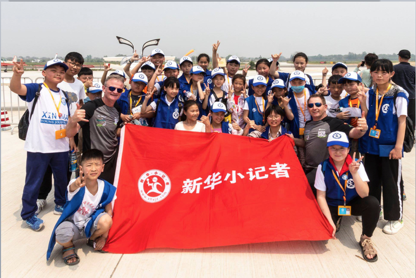 Group of children and two adult men on an airport tarmac holding a red banner with Chinese characters and a logo, posing for a photo.