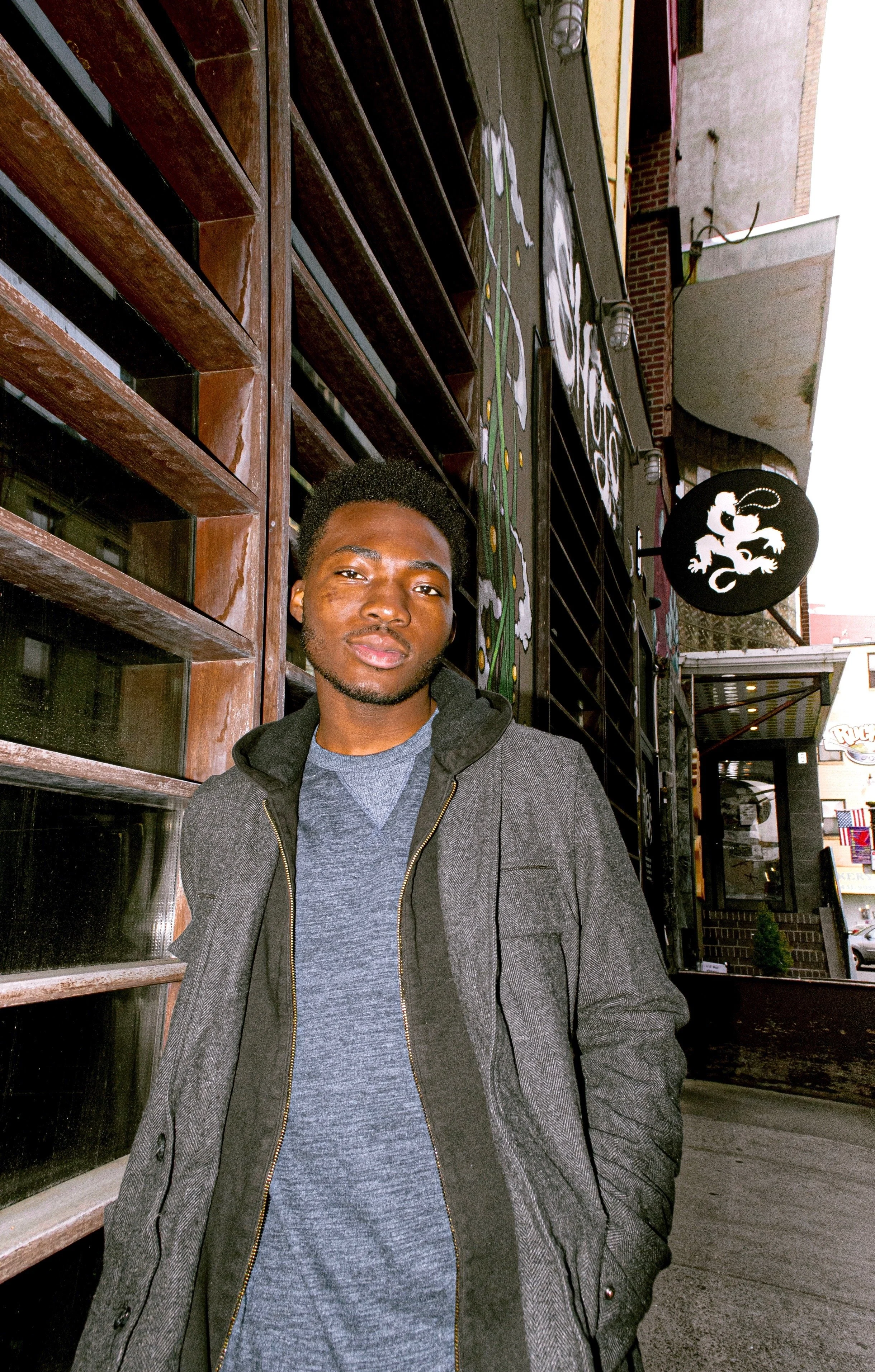 A young man in a gray jacket and blue shirt standing outside a building with wooden and brick elements. The building has a round black sign with a white lizard logo hanging above the entrance.