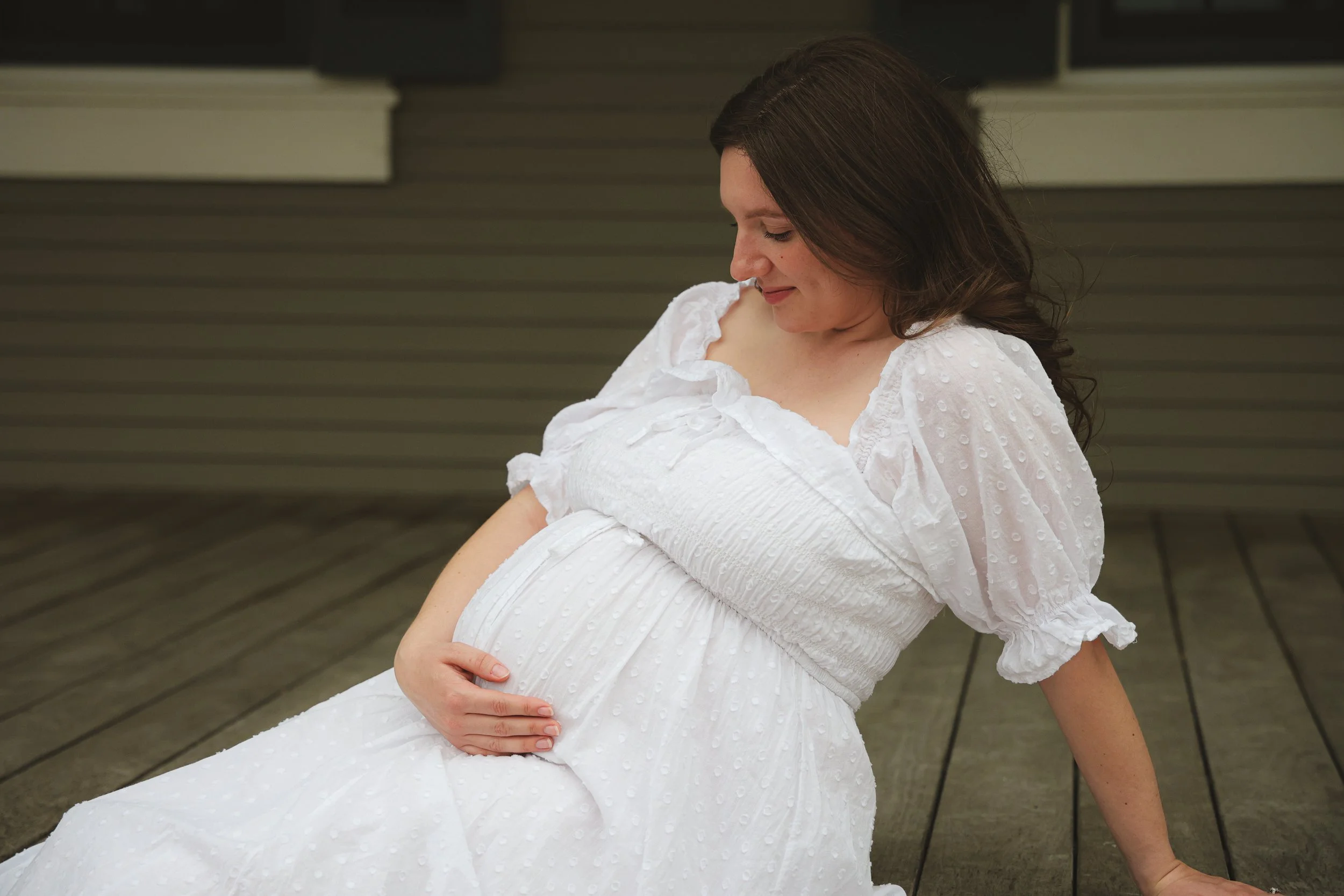 Pregnant woman sitting on wooden floor, gently holding her baby bump, wearing a white dress with textured fabric and puffed sleeves, looking down with a gentle smile.