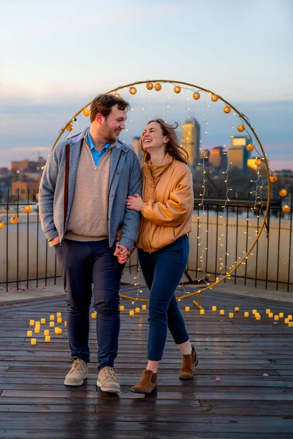 A couple holding hands and smiling at each other on a rooftop with city skyline in the background, decorated with string lights and yellow candles.