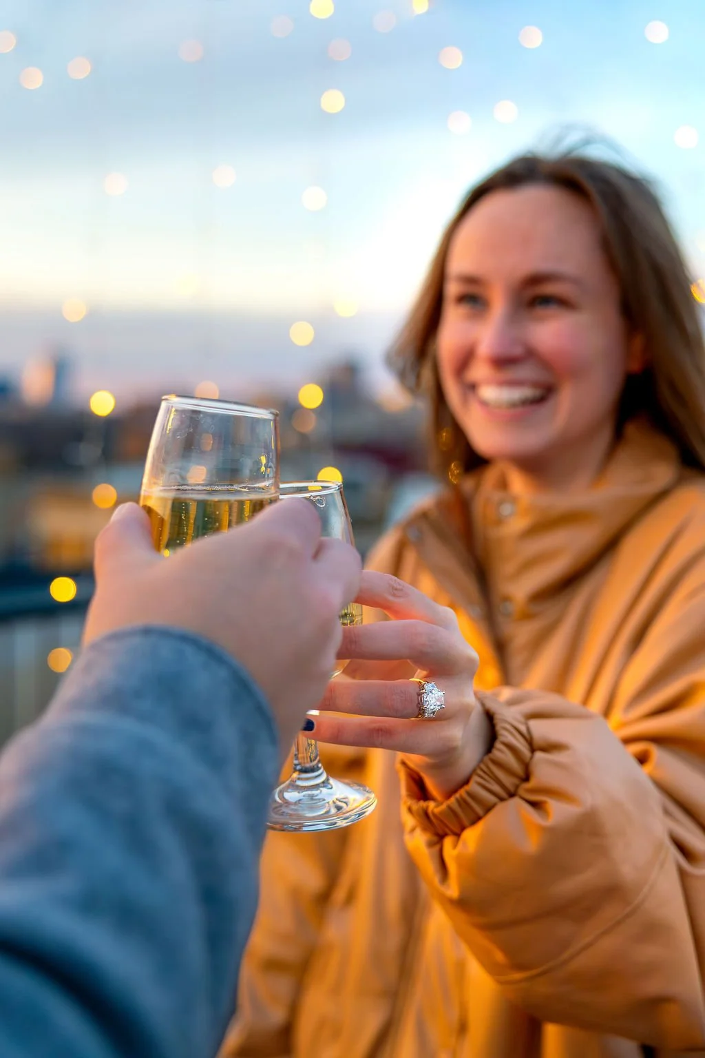 A woman toasting with a glass of champagne against an outdoor cityscape at sunset in Indianapolis, celebrating a proposal, with an engagement ring on her finger.