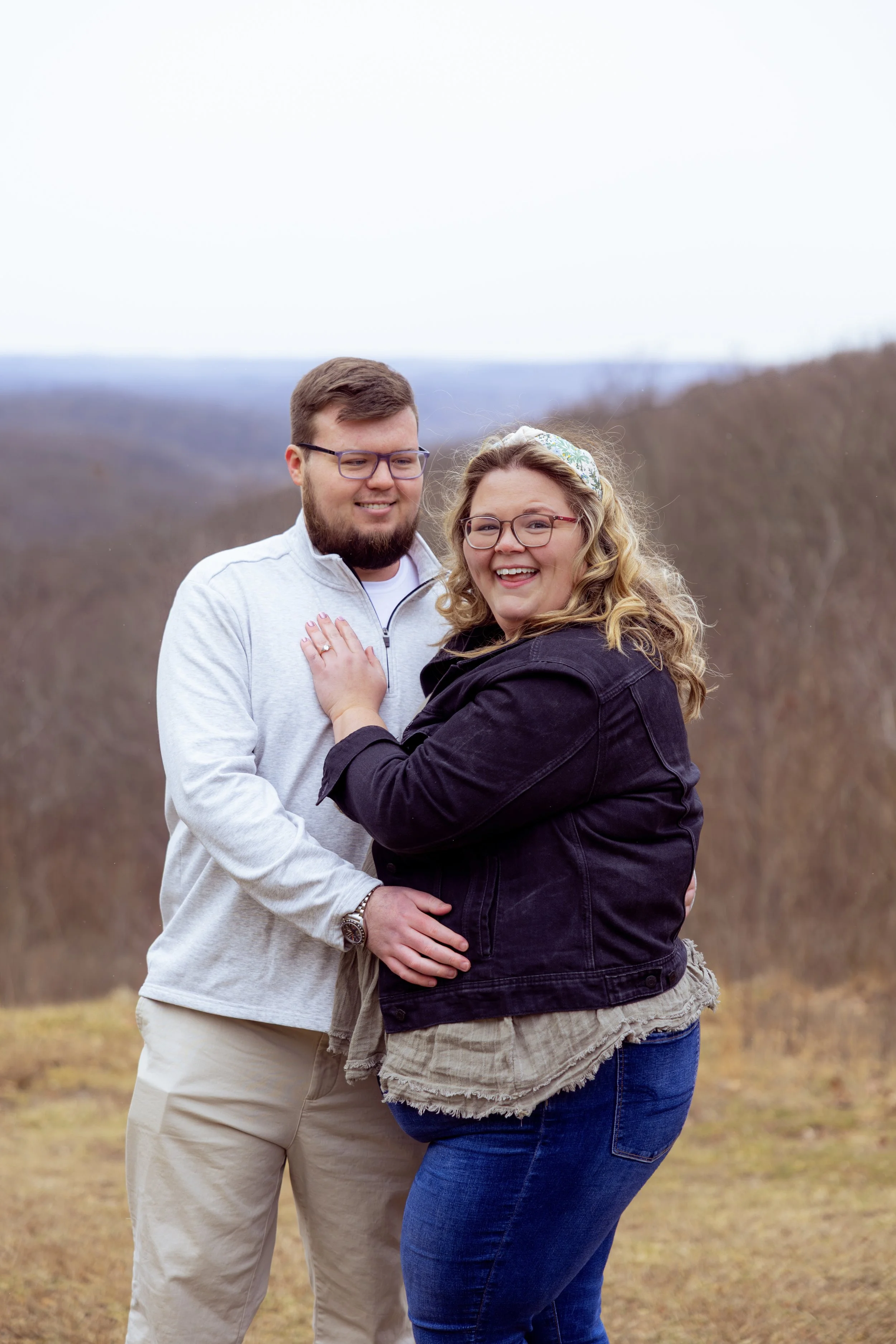 A smiling couple standing outdoors in a hilly landscape, embracing each other and looking happy.