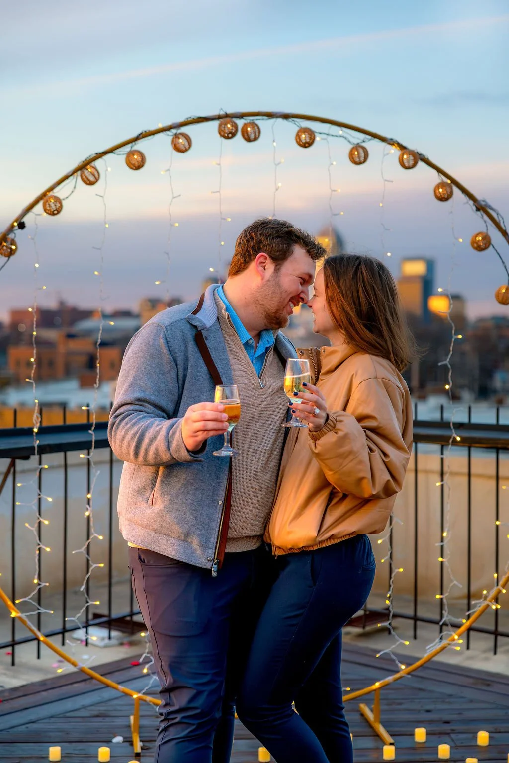 A couple celebrating with glasses of wine on a rooftop at sunset, decorated with a circular string of lights and candles.